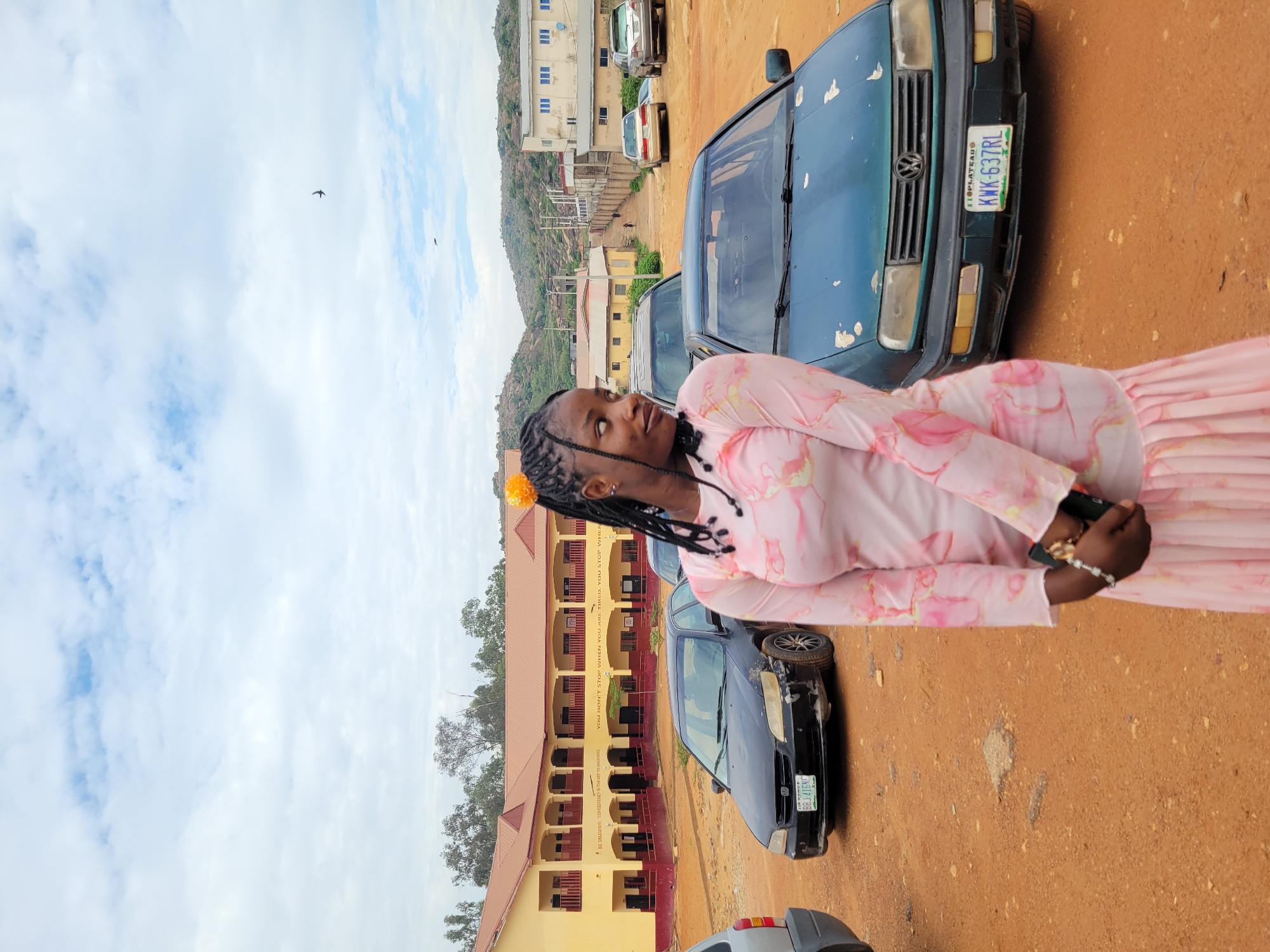 The image depicts an outdoor scene in Gura Top, Nigeria, under an overcast sky with scattered clouds. In the foreground, a young woman with dark braided hair, adorned with a yellow floral accessory, stands facing slightly left with her head turned to the right, looking off-camera. She wears a long-sleeved pink dress with a subtle floral or abstract pattern and holds a dark-colored phone with both hands clasped in front of her, displaying a beaded bracelet on her left wrist. The ground beneath her and extending throughout the midground is reddish-brown laterite, characteristic of unpaved surfaces.
In the midground, several vehicles are parked. To the left, a partial view of a grey/silver vehicle's rear is visible. Immediately behind the woman, a dark-colored Honda sedan with a damaged front bumper is parked, displaying a Nigerian license plate "BBJ-416NF" from Ogun state. To the right of the woman, a dark green Volkswagen sedan, showing signs of paint wear and peeling on the hood, is parked with a Nigerian license plate "KWK-637RL" from Plateau state. Further in the background, an unpaved road ascends a slight incline, flanked by utility poles and wires. Along this road, a light-colored sedan and a silver SUV/sedan are parked.
The background is dominated by a large, two-story, L-shaped building with a yellow facade and a red-brown roof, featuring multiple arched balconies on both levels. Text is visible on the building walls, including "BE DILIGENT: CONSISTENCY IS THE KEY TO GREATNESS" and "YOU DON'T STOP WHEN YOU ARE TIRED... YOU STOP WHEN YOU ARE DONE." This structure appears to be an educational or institutional building. To the right of this building, other structures are visible, including a white two-story building with blue-framed windows and another yellow building. The far background reveals rocky, vegetated hills under the cloudy sky. Two dark, bird-like silhouettes are discernible in the upper right portion of the sky.