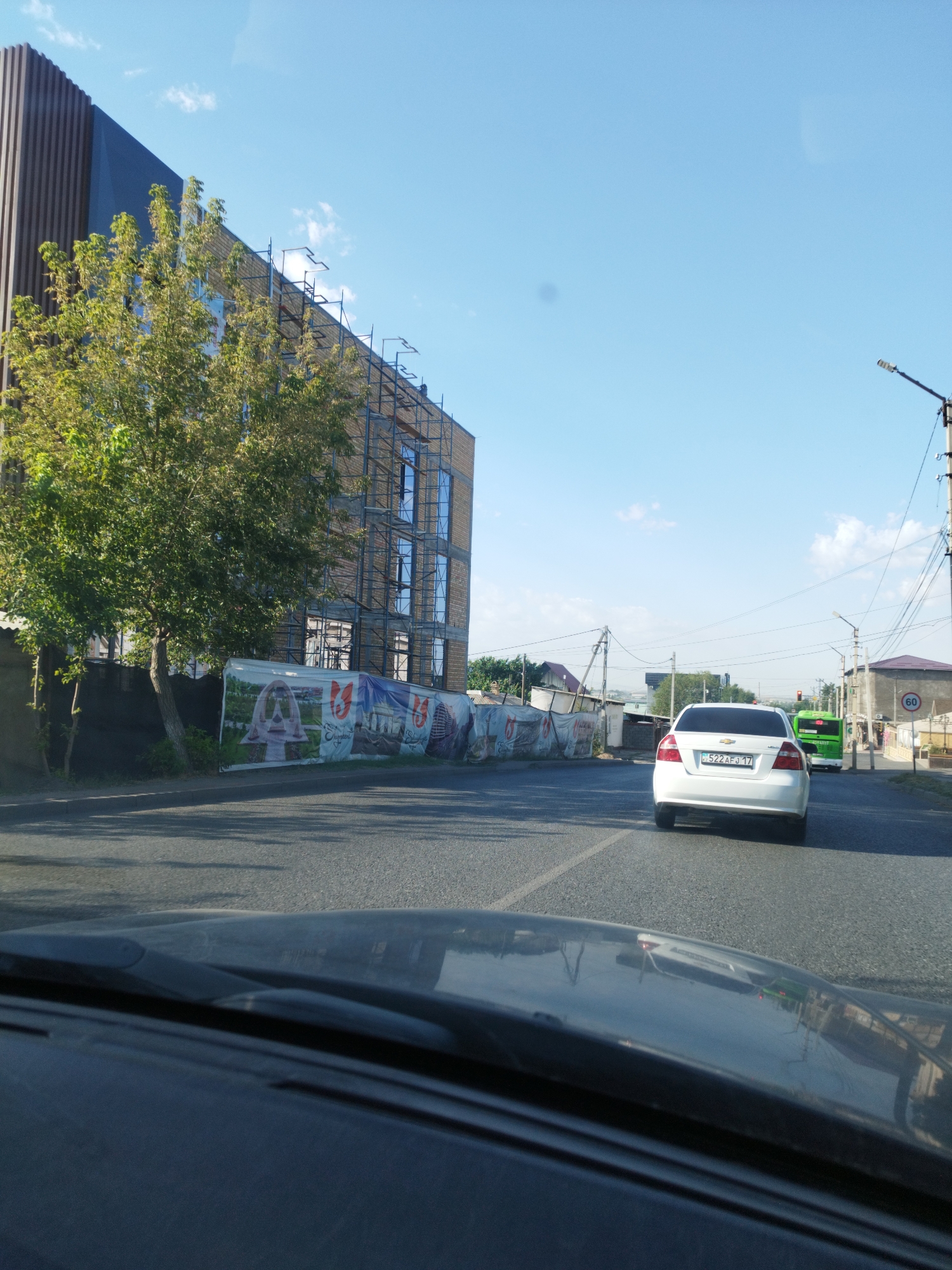 This is a street-level, eye-level shot taken from a vehicle, looking forward on a sunny day in Shymkent, Kazakhstan. The scene depicts a street with two lanes of asphalt. On the left side of the street, there is a multi-story building under construction, partially covered with scaffolding. A large banner advertising the building's future appearance is visible in front of it. A mature tree with green foliage stands in front of the building. On the right side of the street, a white car with license plate "522 AFJ 17" is driving away from the camera, followed by a green bus. Further down the street, a traffic light is visible, and a speed limit sign of "60" is posted. Power lines and streetlights are present along the street. The sky is clear and blue with scattered white clouds.