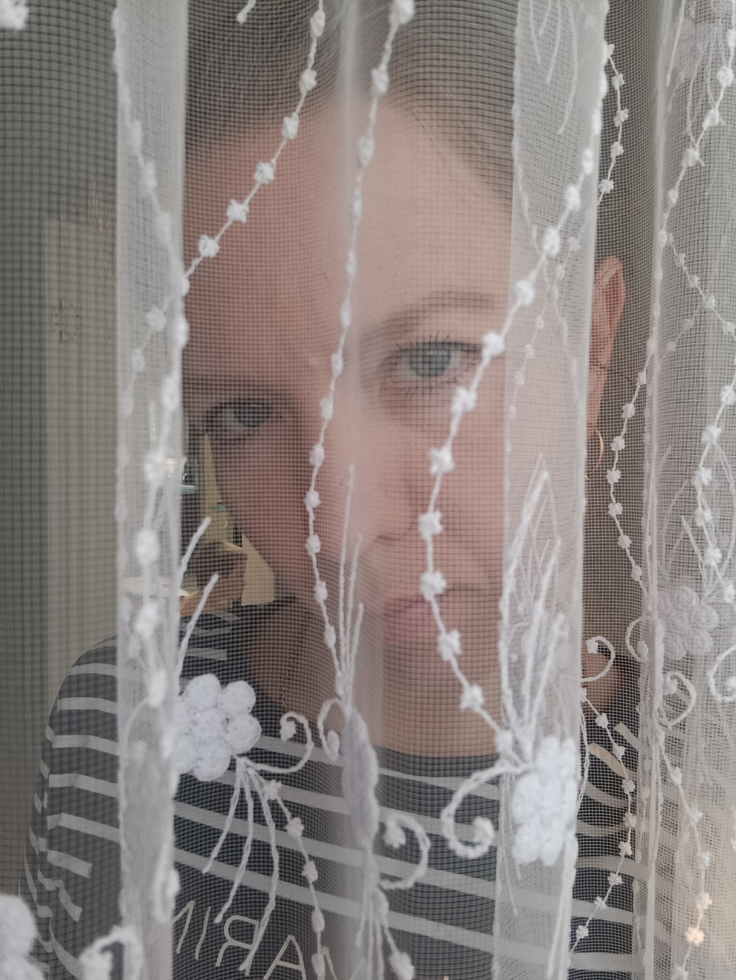 An individual, likely a young adult or child, is partially visible behind a sheer white curtain. Their face, obscured by the curtain's mesh and decorative elements, features one prominent blue or green eye gazing directly at the viewer. Parts of their nose and mouth are also discernible, along with their ear on the right side of the frame. The person is wearing a striped top, possibly black and white or dark and light grey.
The white curtain, which dominates the foreground, is intricately patterned with embroidered or crocheted floral designs. These patterns are in clear focus, contrasting with the slightly softer focus on the person behind. The setting appears to be indoors, likely near a window, with ambient daylight providing illumination. The background beyond the person is indistinct, suggesting a typical room environment.
The activity depicted is that of peeking or looking out from behind a curtain, conveying a sense of curiosity, shyness, or playful engagement. Visible text on the person's striped shirt, seen in reverse through the translucent curtain, appears to spell "AIRA" or a similar word. The scene is set in Chaikovsky, Russia.
