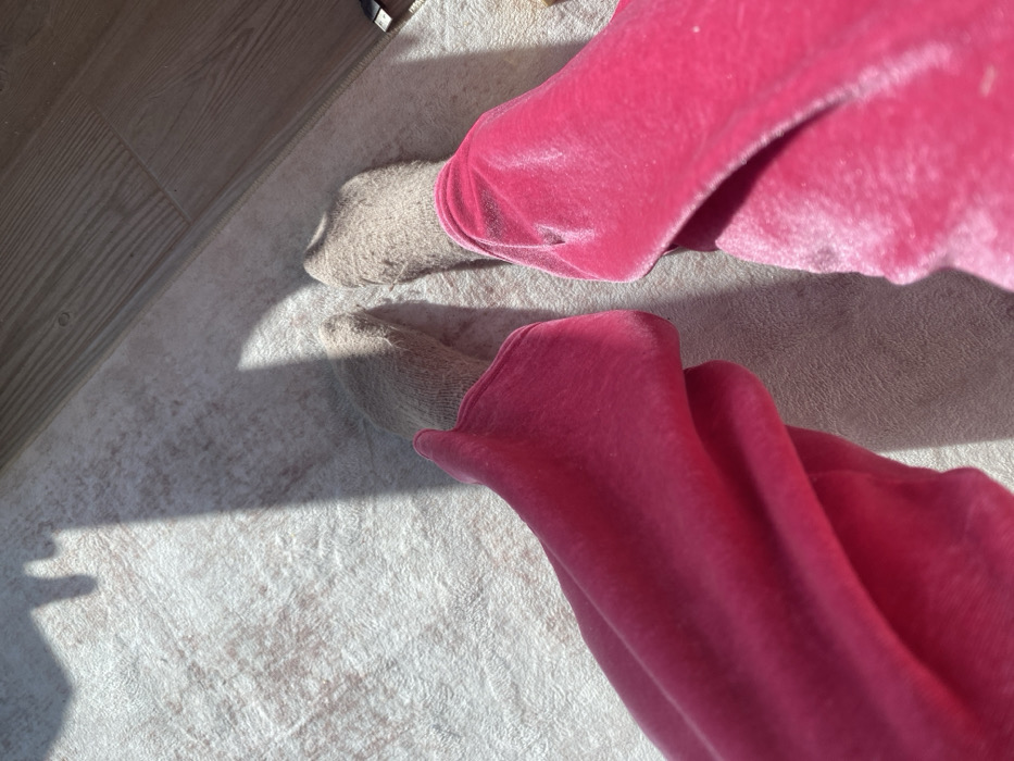 The image displays an overhead view of a person's lower legs and feet. The individual is wearing bright pink, likely velour or velvet, trousers that are gathered around the ankles. Both feet are covered in light brown knitted socks. The feet and socks are positioned on a light-colored, textured rug or carpet. A strip of light-toned wooden flooring is visible along the upper right edge of the frame, adjacent to the rug. Prominent, elongated shadows are cast across the rug, indicative of direct, strong lighting. The scene provides no discernible details to confirm the specific location as Miami, United States of America.