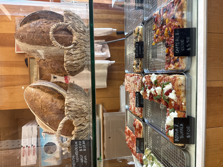 The image displays a food counter inside an establishment in New York, United States of America, likely named 'Impasto' based on visible branding. The scene is viewed through a clear glass display case.
On an upper glass shelf, two large, oblong loaves of dark-crusted sourdough bread are presented, each resting in a light brown, woven basket with a handle. A small black sign on the left indicates 'HOUSE SOURDOUGH' prices: 'HALF $7.00' and 'FULL $12.00'. Behind this shelf, stacked white pizza boxes branded 'IMPASTO' are visible on the left, and a wooden paneled wall forms the background. A reflection shows a partial view of a person wearing a white shirt with red 'IMPASTO' text.
On a lower shelf, a variety of rectangular pizza slices are displayed on metal racks within metal trays. From left to right, visible pizza types include: 'PEPPERONI' ($5.50), 'MARGHERITA' ($4.50), and 'SALSICCIA & FUNGHI' ($6.50). A partial view of a green-topped pizza and a 'PORCHE POTATO' pizza is also present. Below these, an L-shaped portion of 'ROSSA & BUFALA' pizza, topped with white cheese dollops and basil leaves, is priced at '$7.00'. Adjacent to it is a rectangular 'CAPPERI & OLIVE' pizza topped with colorful chopped vegetables, priced at '$5.50'. The countertop beneath the display is white, with a wooden base below it.