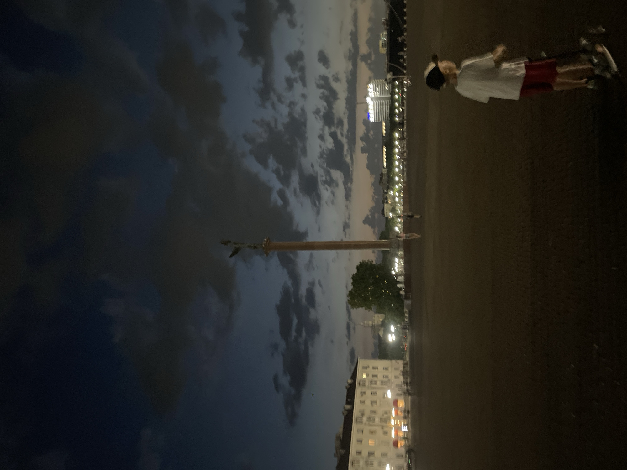 This is a medium-shot, eye-level, outdoor photograph taken at dusk or early night. The scene depicts an open, paved area with a prominent monument on the right side, a large building on the left, and a cityscape in the background.
In the foreground, a young boy, appearing to be Caucasian, is standing to the right of the frame, facing towards the left. He is wearing a white t-shirt, red shorts, a baseball cap, and what appear to be sneakers. He is standing next to a scooter, with his hands resting on the handlebars. His body is angled slightly away from the viewer.
The middle ground features a vast, dark paved surface, possibly a plaza or square. Further back, on the left side of the image, is a large, light-colored building with illuminated windows, suggesting it is occupied or in use. Adjacent to this building, a tall, slender monument with a statue at its apex rises into the sky. Behind the monument, trees are visible. Across the expanse, a line of streetlights illuminates the distance, with more buildings forming the cityscape. The sky is dark blue, with scattered clouds that are tinged with lighter colors from the fading daylight or artificial lights. The overall lighting is dim, creating a twilight atmosphere.
The location is identified as Verkhnyaya Stavropol'ka, Russia.