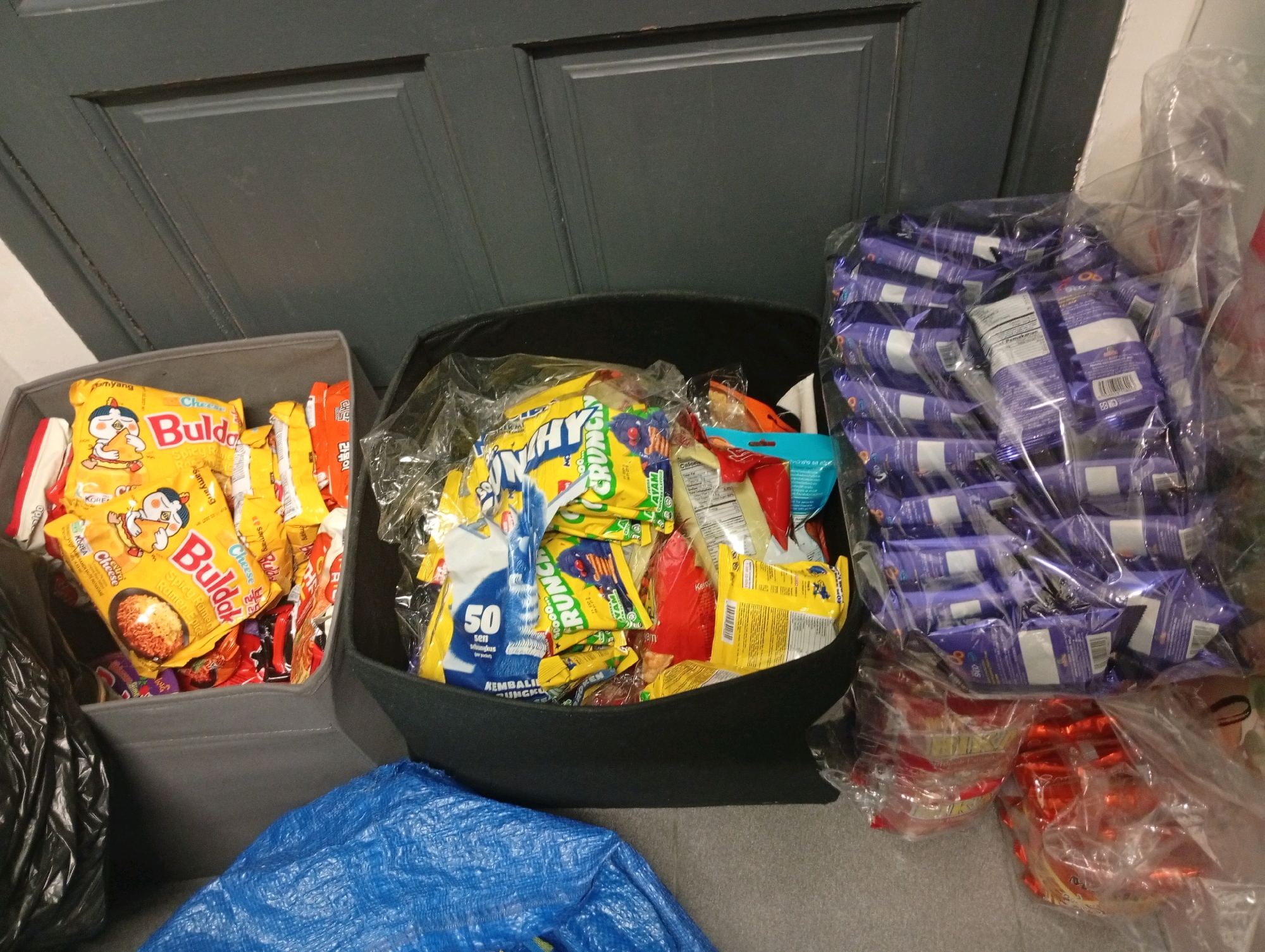 The image displays an assortment of food items, predominantly packaged snacks, within a residential setting. Two grey fabric bins are visible, one filled with brightly colored instant noodle packets, notably "Buladak Cheese Spicy Ramen" and other Samyang brand products. Adjacent to this bin is a black fabric bin, overflowing with a variety of snack bags, including items with blue and yellow packaging featuring text that appears to be in Malay, such as "50 sen." To the right, several plastic-wrapped packages of small, rectangular food items with purple and white branding are stacked. These are consistent with individually wrapped snacks or candies. Beneath these, a collection of red and silver wrapped items, possibly candies or biscuits, are also present, contained within plastic. A large blue plastic sheet is spread on the floor in the foreground, and a black plastic bag is situated to the left. The background features a dark-colored door with paneling. The overall impression is that of a collection of food items, likely groceries or supplies, stored in a dwelling. The presence of items with Malay text suggests the location is within Malaysia, potentially in an area like the Selayang Municipal Council.