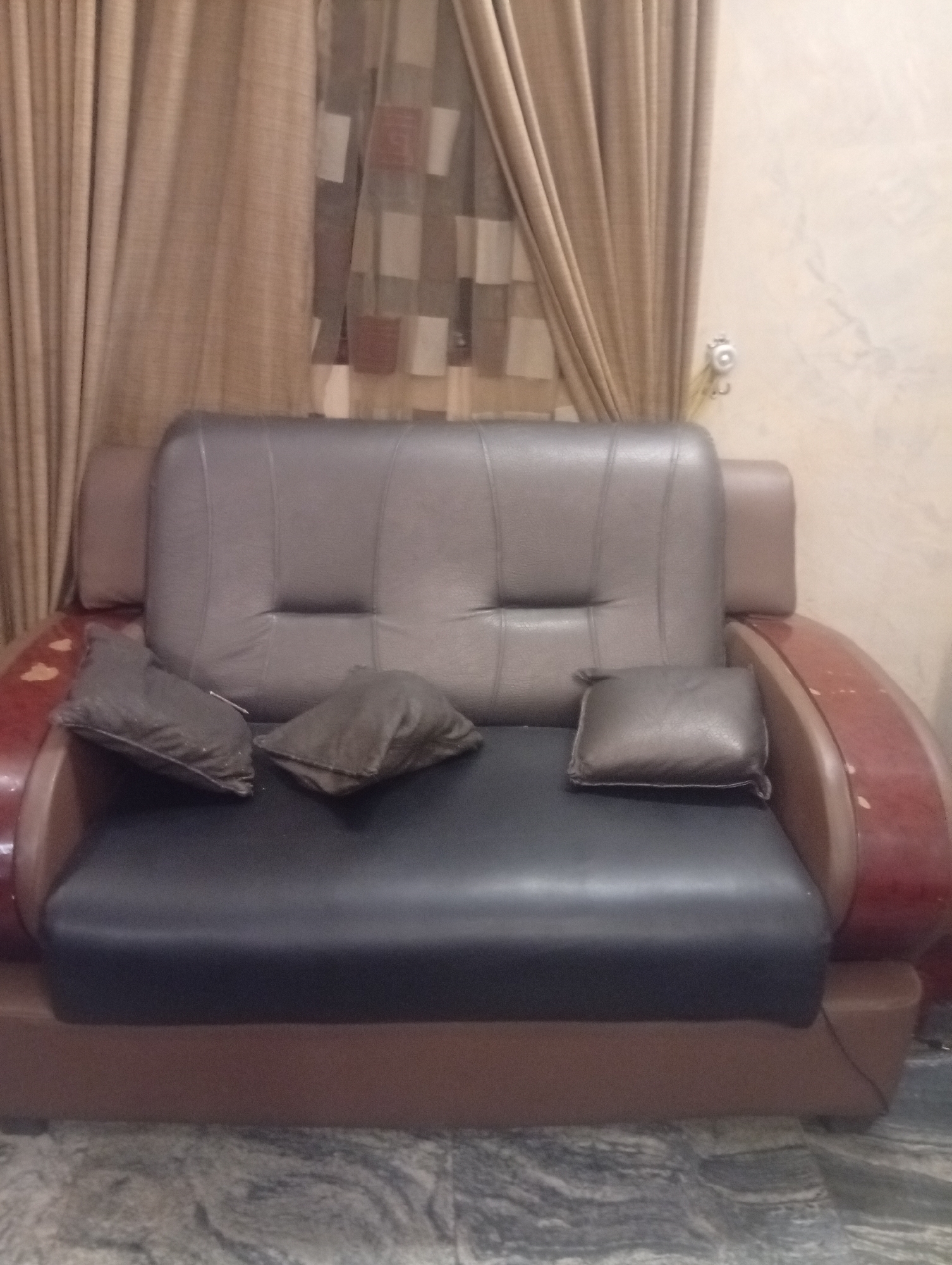 The image depicts a brown and black leather sofa with three brown pillows on it, set against a background of beige curtains and a beige wall. The sofa shows signs of wear and tear, notably on the wooden armrests. The scene is indoors, with a glimpse of a marble floor at the bottom of the frame. The location is identified as Aviation Quarters, Nigeria. There are no people visible in the image.