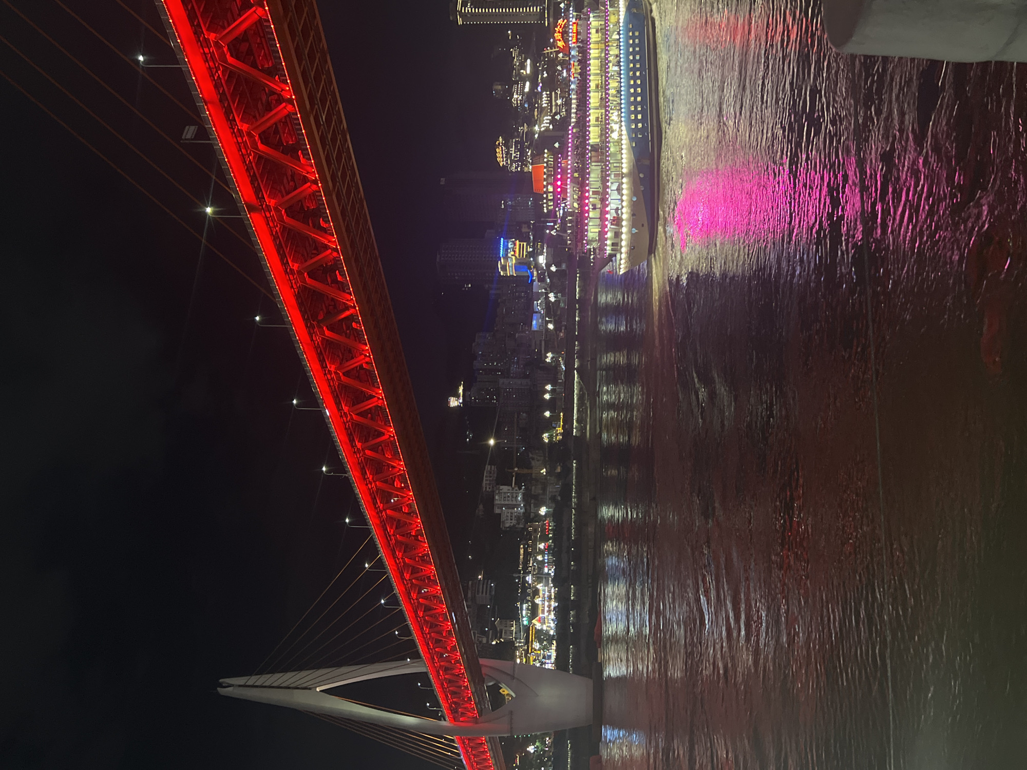 An illuminated bridge spans a dark river at night. The bridge's structure is a vibrant red, casting a bright glow. Cables extend from a white support pillar on the left, connecting to the bridge's deck. In the river, a multi-story boat with numerous windows and decorative lights is moving, its pink and white illumination reflecting on the water's surface. The background shows a cityscape with buildings of varying heights, also lit up at night, suggesting a dense urban environment. The water appears slightly choppy, reflecting the surrounding lights in streaks and patterns. The sky above is dark and clear. This scene is captured in the Yuzhong District of China, likely a waterfront area known for its illuminated bridges and bustling nightlife.