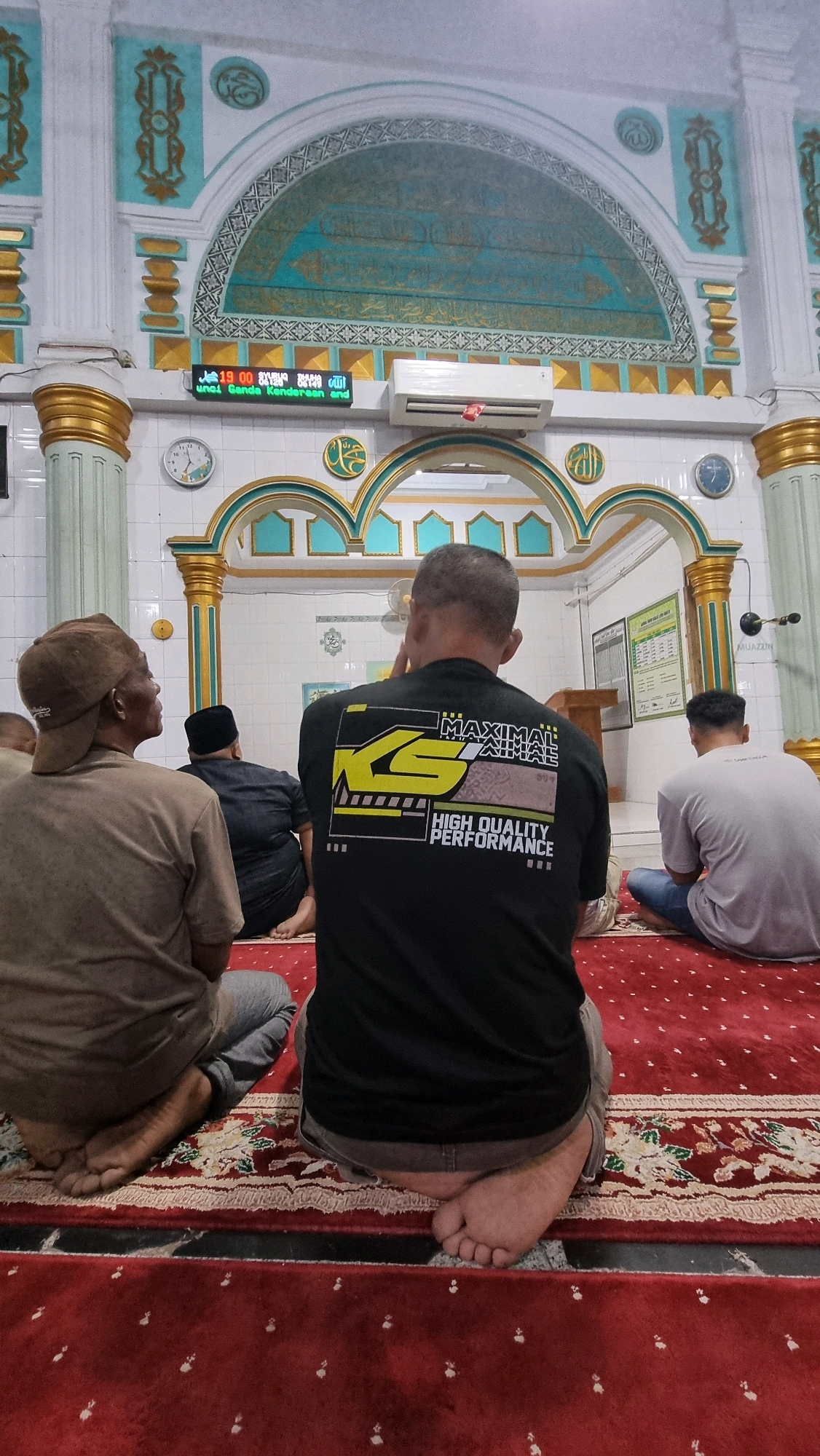 In Puloara, Indonesia, inside a mosque, several individuals are seated on a red carpet. The mosque features white walls with ornate teal and gold architectural details. Above, Islamic calligraphy is visible. In the center of the image, a person wearing a black t-shirt with a yellow and black graphic design is seen from behind. To their left, another person in a brown cap is also seated. To the right, a person in a light gray t-shirt is visible. A digital display showing the time and a clock are mounted on the wall. An air conditioning unit is situated above the digital display. The scene depicts a moment of prayer or reflection within the mosque.