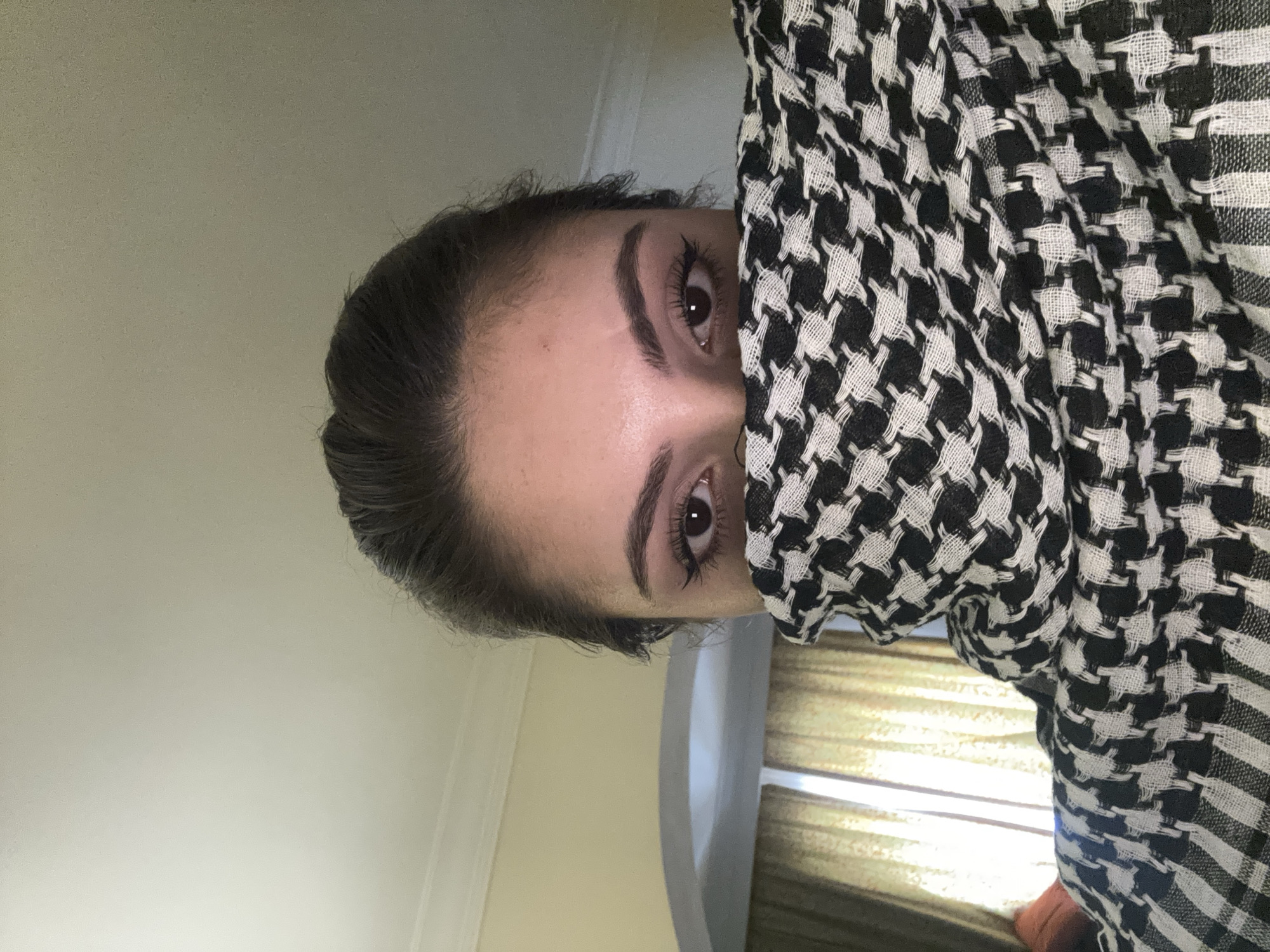 A person is shown from the forehead to the lower part of the nose with the right side of the face covered by a black and white patterned cloth. The person has dark hair pulled back and visible dark eyebrows and eyes. The background appears to be an interior setting with a light-colored wall. A window with tan-colored curtains is slightly visible to the person's left side. The location is unknown.