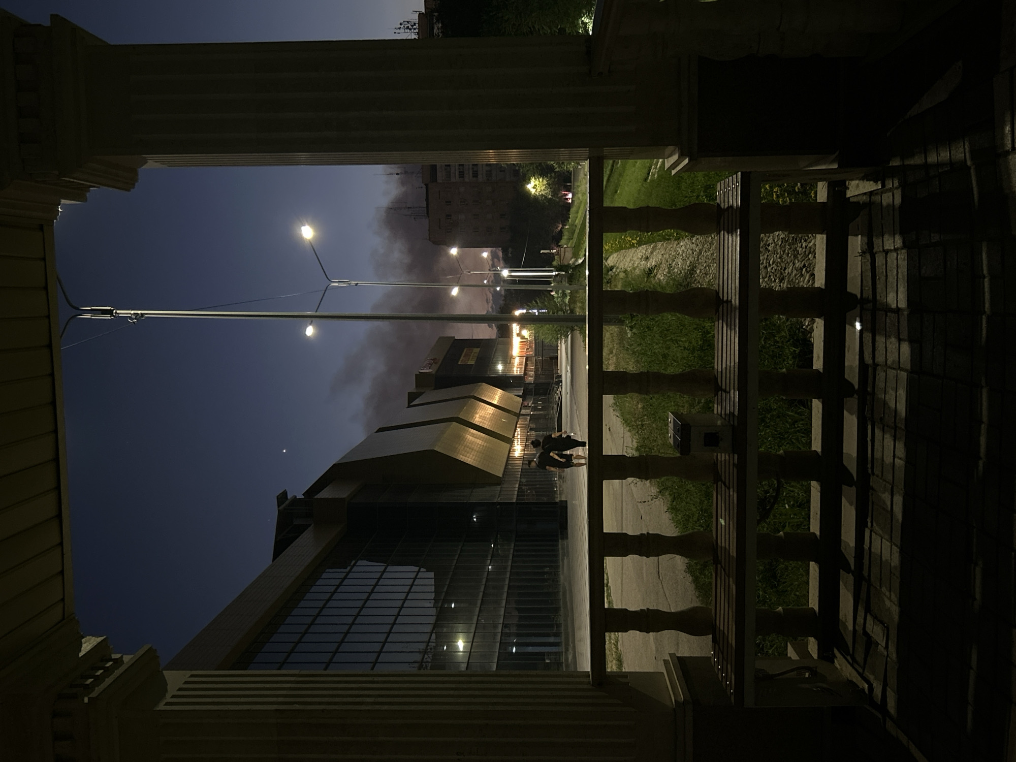 The image, taken at dusk in Taraz city administration, Kazakhstan, depicts an exterior scene from a covered porch with ornate pillars. In the foreground, a wooden bench with a metallic solar-powered light fixture sits on a brick-paved area, casting long shadows. A white stone balustrade separates the porch from a grassy area. Beyond this, a modern building with a glass facade and gold accents is visible, with two figures walking in front of it. Further in the background, streetlights illuminate the twilight sky, and smoke plumes rise from an area with apartment buildings.
