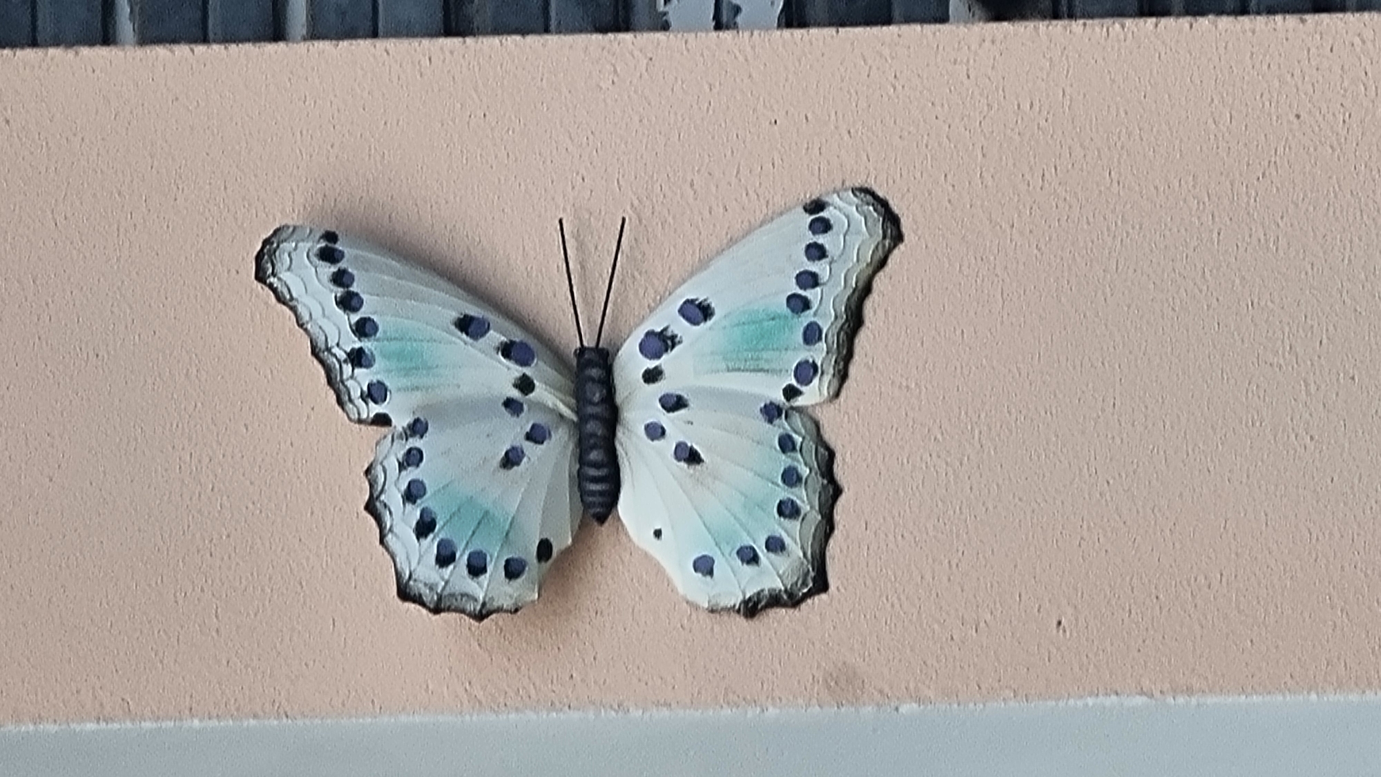 A decorative butterfly sculpture is affixed to a textured, light pink wall. The butterfly has light blue and white wings with numerous dark blue spots. The butterfly's body is black and segmented, and two thin black antennae extend from its head. The wall appears to be an exterior surface, as evidenced by a portion of a dark, possibly brick, structure visible on the right edge of the image. The surrounding environment is not clearly discernible beyond the wall and the adjacent structure. This scene is located in Sant Joan les Fonts, Spain.