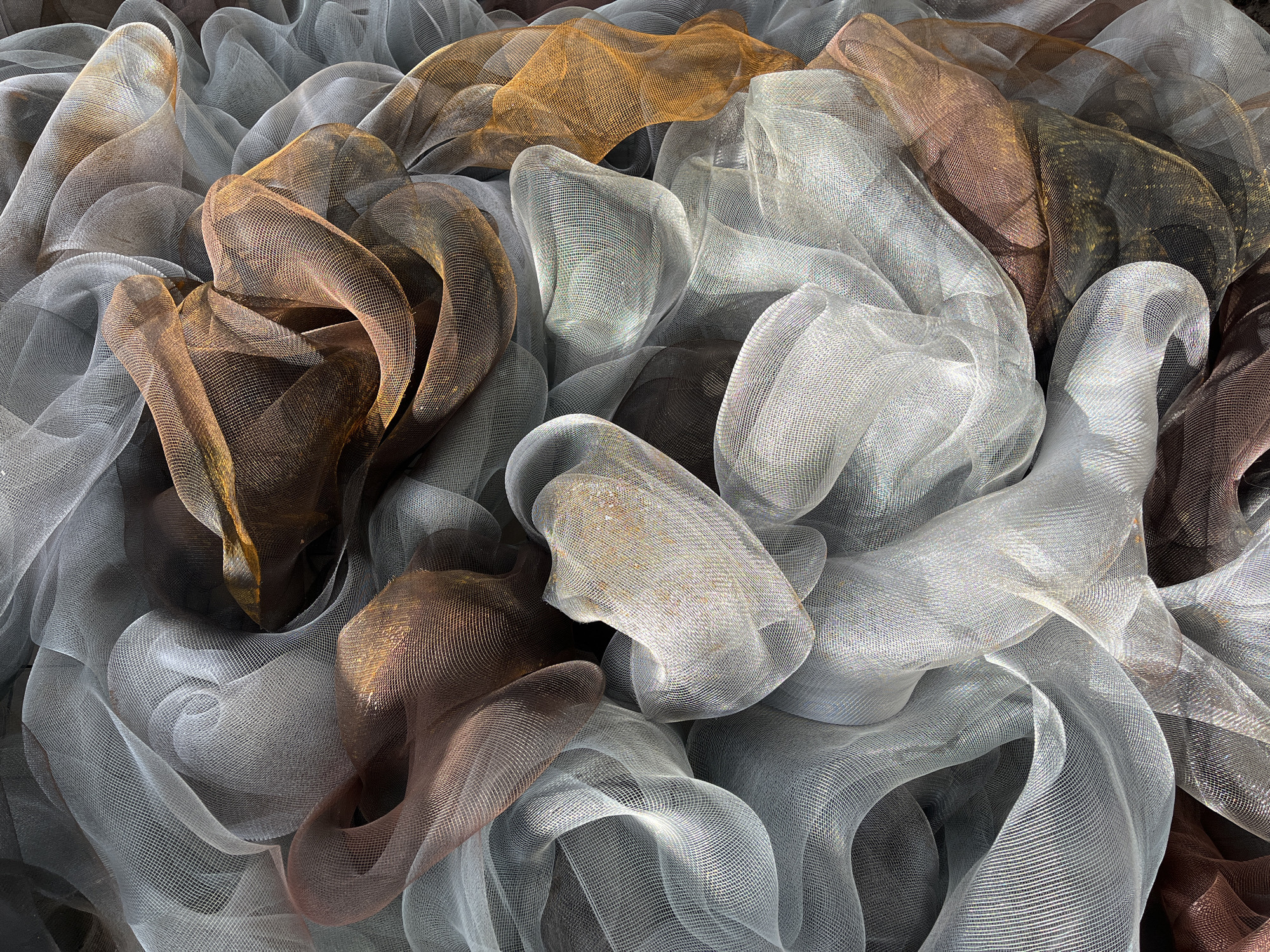 The image shows a close-up, overhead view of a pile of metallic netting fabric in silver, gold, and brown hues. The fabric is arranged in loose, flowing folds and curves, creating a textured and layered effect. The netting is translucent, allowing light to pass through and revealing the varying colors and depths of the pile. There are no people or recognizable objects in the immediate vicinity. The environment is undefined, with the focus entirely on the arrangement of the metallic fabric. The city of Krasnoyarsk, Russia, is not apparent in the visual information.