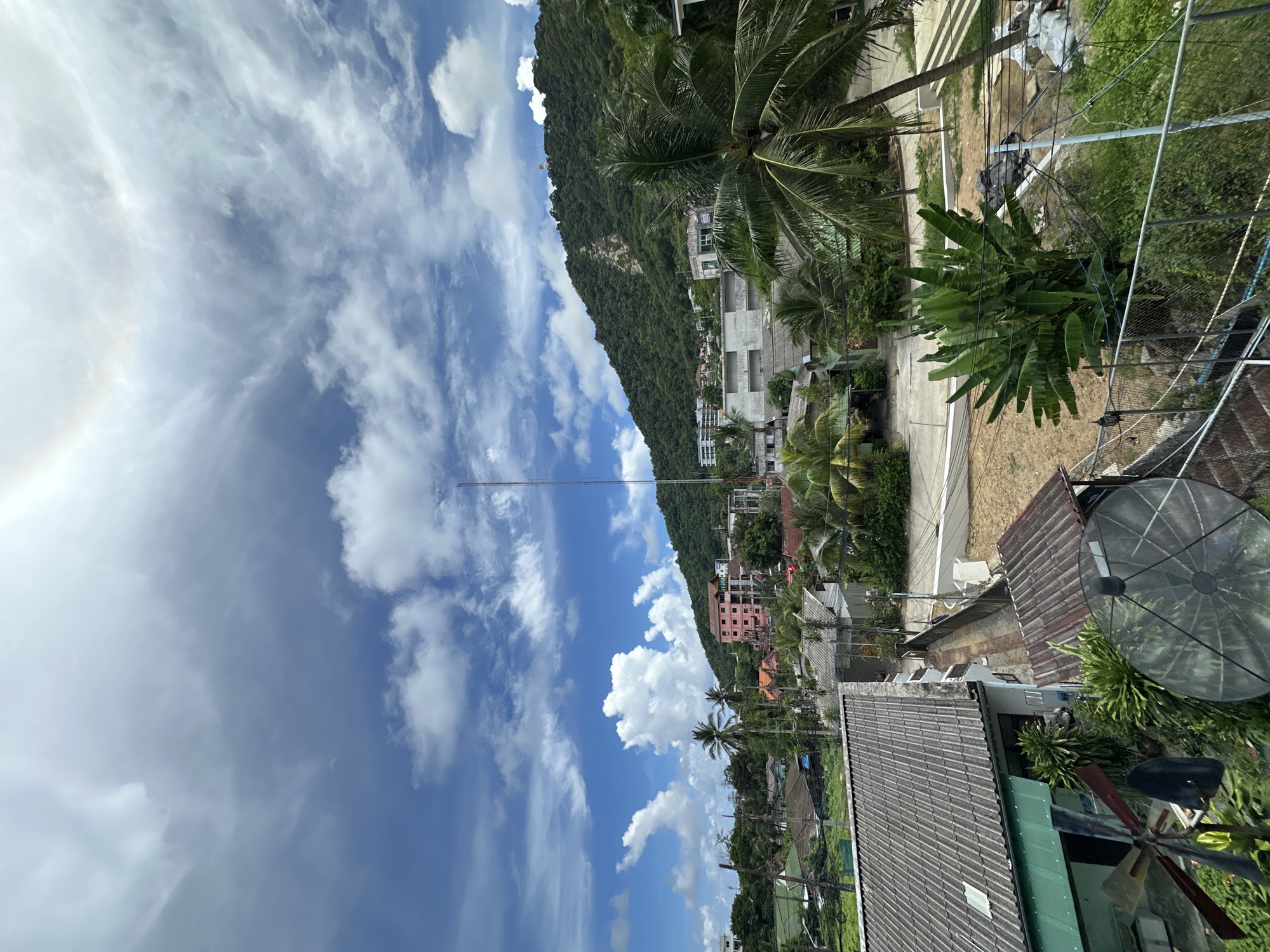 The image depicts a tropical scene in Karon, Thailand, with a steep, tree-covered mountain dominating the left side of the frame. The sky is a vibrant blue, filled with scattered white clouds, suggesting a partly cloudy day. A thin, tall antenna or mast rises from the base of the mountain, reaching towards the sky.
Below the mountain, a cluster of buildings is visible, including a multi-story pink building and several other structures with white or grey facades, interspersed with lush green palm trees and other tropical vegetation. The architecture suggests a developed area, possibly a town or resort.
In the foreground, the roofs of several houses are visible. One house has a gray tiled roof, and another has a darker, corrugated metal roof. A large satellite dish is mounted on the roof of one of the buildings, pointing towards the sky. Various wires and electrical poles crisscross the scene, indicating the presence of infrastructure. The overall impression is one of a coastal or island community nestled against a mountainous backdrop. The time of day appears to be daytime, likely late morning or early afternoon, given the brightness of the sunlight and the shadows. No people are explicitly visible.