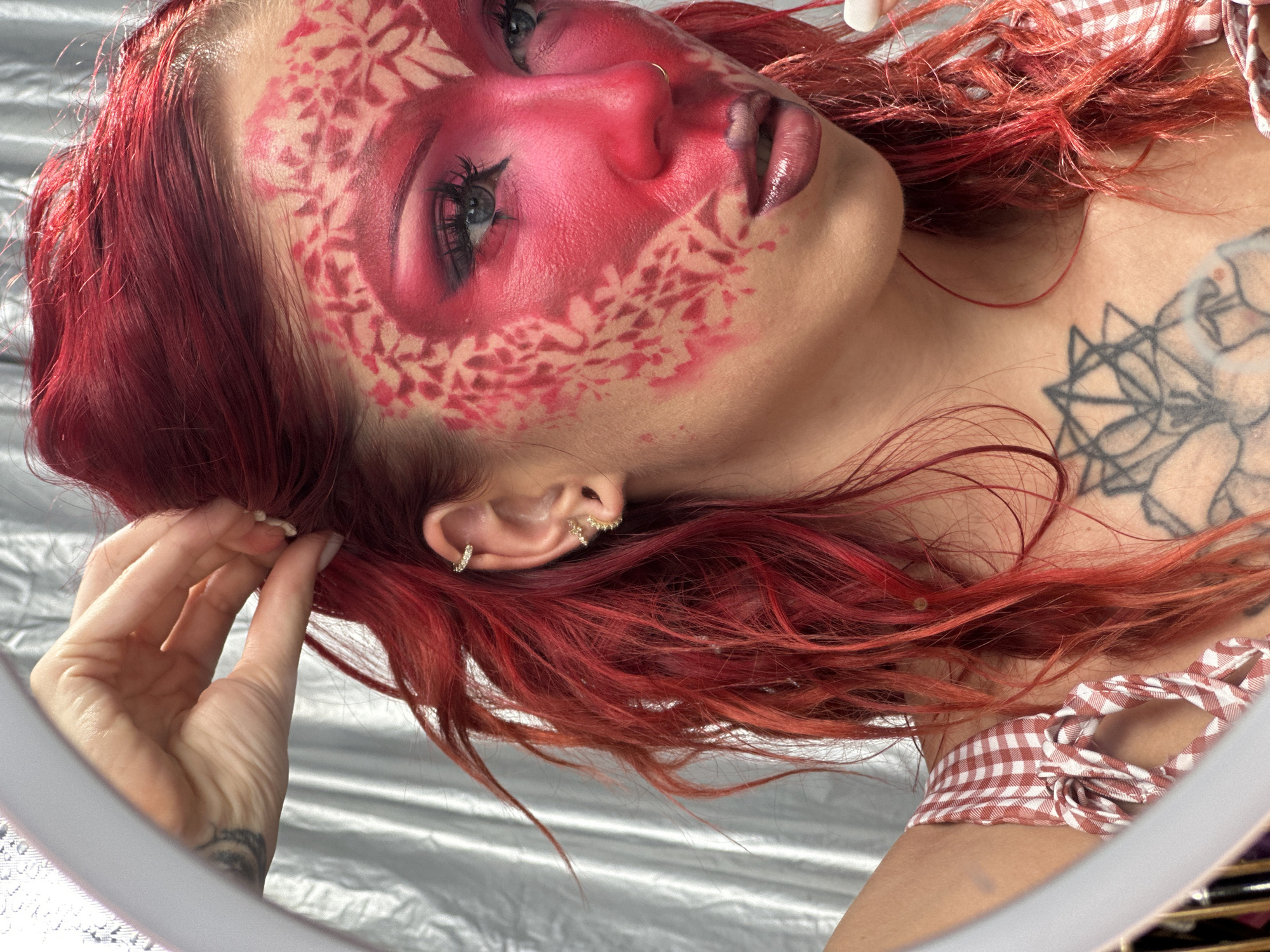 This media file is a close-up, high-angle shot of a person with vibrant red hair. The subject is lying down, with their head tilted upwards, looking towards the camera. Their face is adorned with striking red and pink face paint, featuring a floral or patterned stencil design across their cheeks and forehead. Their eyes are open, with dramatic eye makeup, and they have a nose ring. Their lips are glossy and appear to be a dark, deep red or purple shade.
The person is wearing a top with a red and white checkered pattern, tied at the front. A tattoo with intricate linework is visible on their upper chest and shoulder. Several small, sparkling earrings are visible in their ear. The person's right hand is brought up towards their face, with their fingers gently touching their hair near their ear.
The background appears to be a textured, metallic silver surface, possibly fabric or a reflective material. The overall mood is artistic and stylized, emphasizing the elaborate makeup and vibrant colors. There is no visible text in the image. The provided location context of "North Kuta, Indonesia" does not offer any specific cues within the visual content of the image itself.