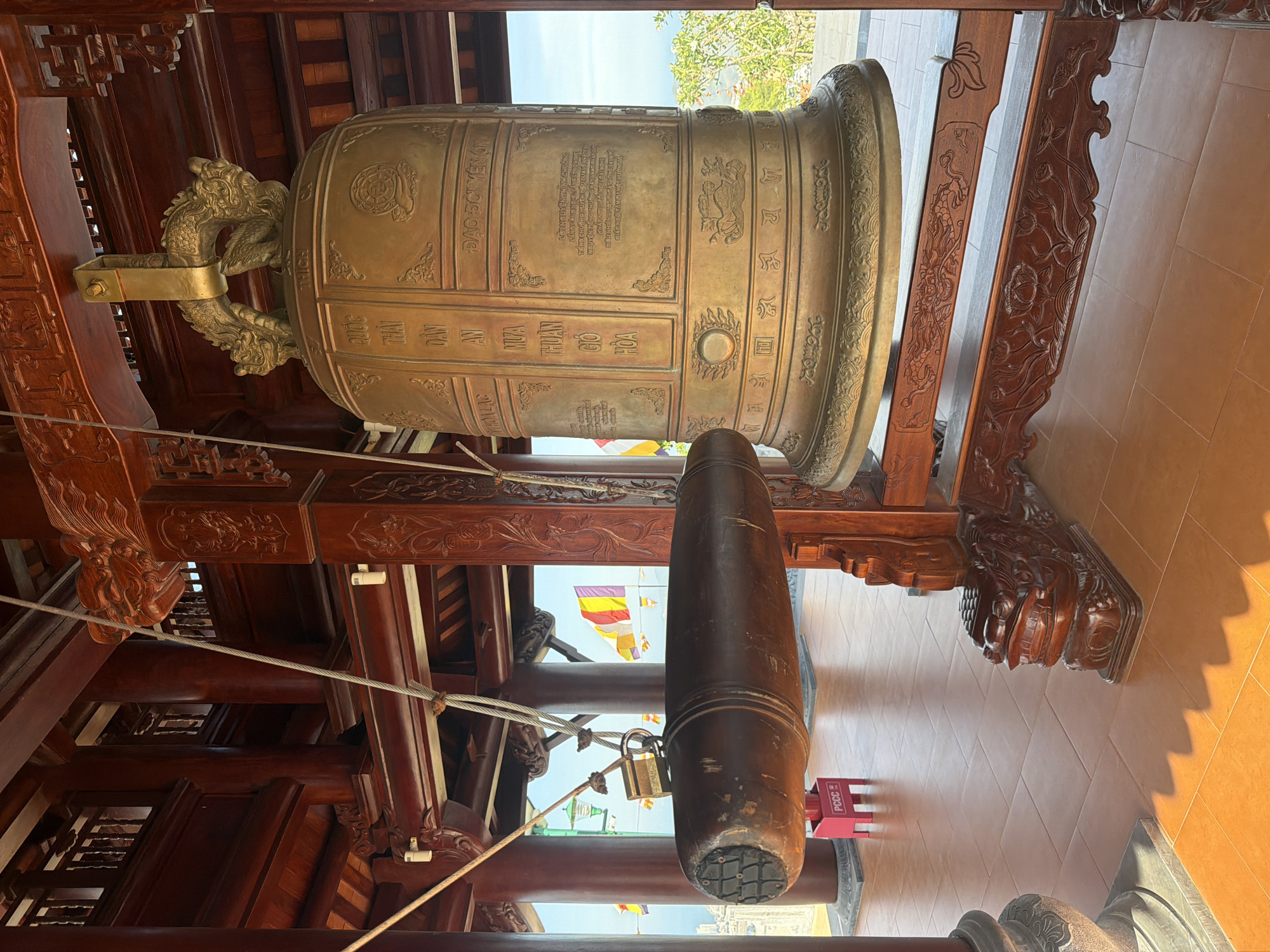 The image captures an interior view featuring a large bronze bell and a wooden striker, situated presumably inside a temple or similar structure in Nam Nha Trang Ward, Vietnam. The bell is adorned with intricate carvings, including a dragon handle and rectangular panels with inscriptions in Vietnamese, some of which are partially legible. The text contains religious or philosophical content, possibly prayers or dedications. The bell is suspended from an elaborately carved wooden frame, indicating a traditional architectural style. Hanging beneath the bell is a thick wooden striker, also suspended from the frame with metal hardware and ropes. The surroundings include tiled flooring and glimpses of the outside, revealing flags and foliage. The overall scene suggests a sacred space designed for contemplation and ritual practices.