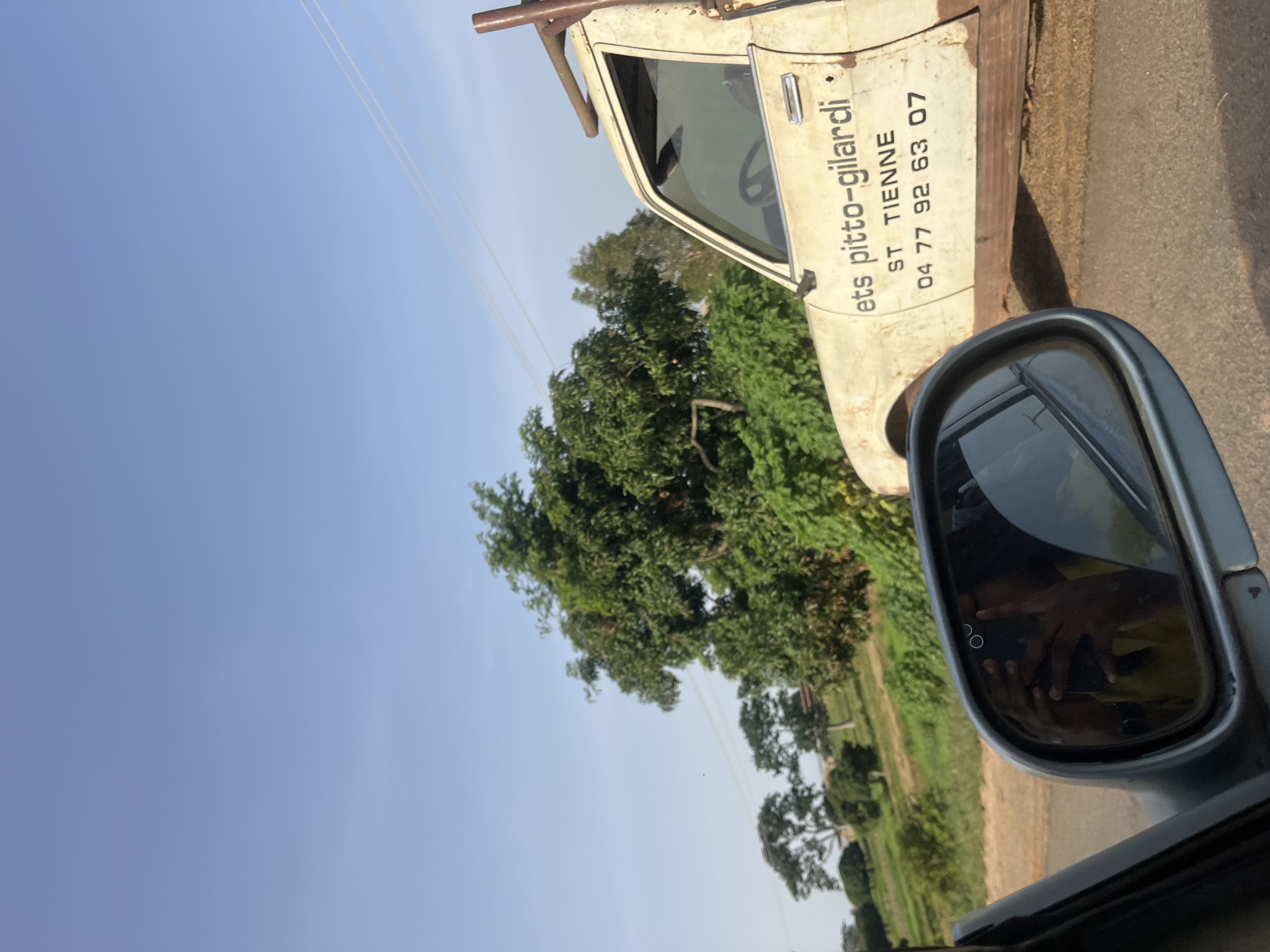 The image shows a side view of an old, weathered white truck with text on its door that reads "ets pitto-gilardi", "ST TIENNE", and "04 77 92 63 07". The truck appears to be parked on a dirt road with a grassy embankment and trees in the background. A side mirror of another vehicle is in the foreground, reflecting the sky and a person holding a phone. The sky is clear and blue. The environment suggests a rural or semi-rural setting, consistent with the outskirts of Ladura Foron, Nigeria.