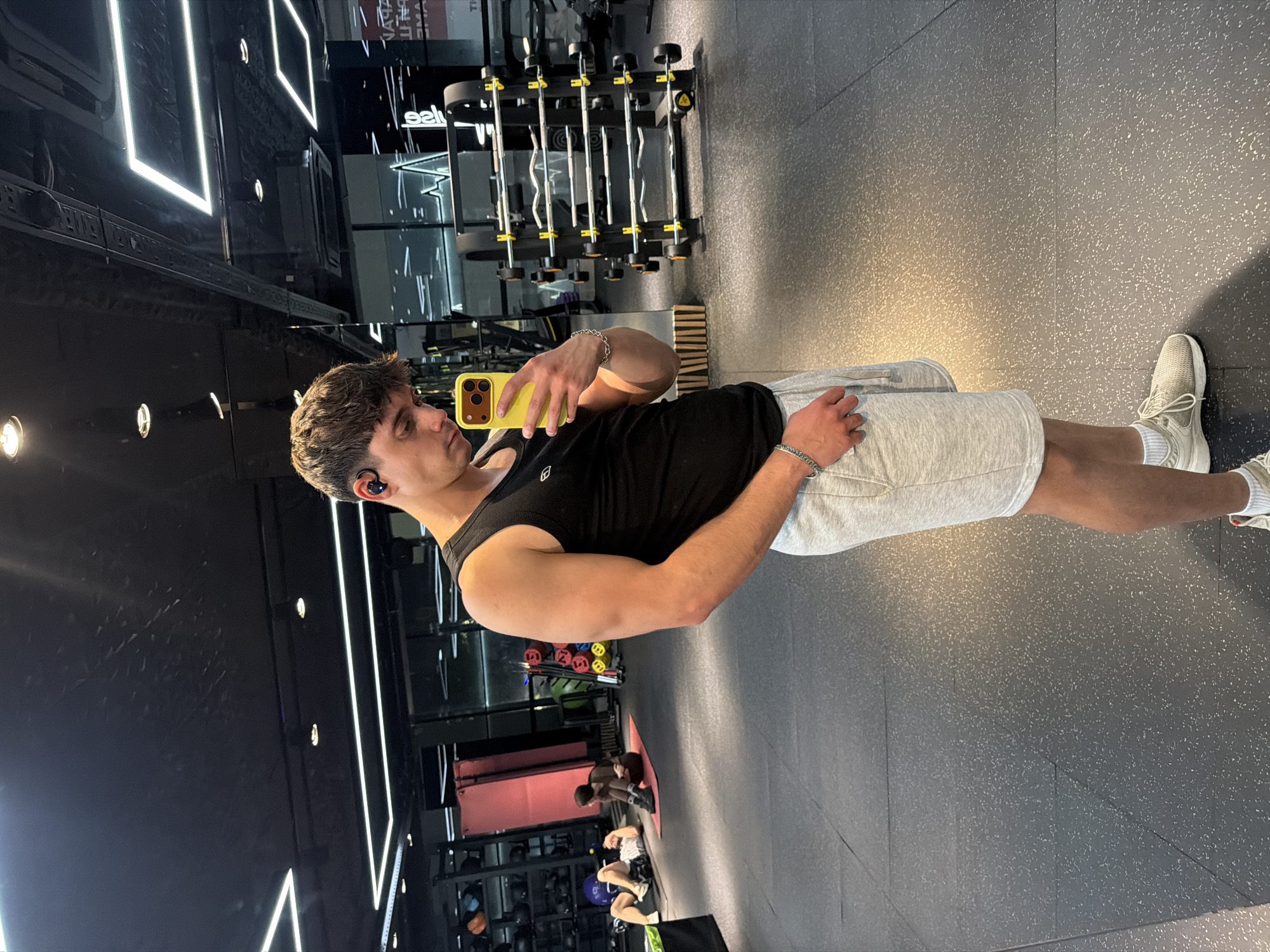 A man is taking a mirror selfie at a gym. He is wearing a black tank top and gray shorts. The floor is dark gray with white flecks. In the background, gym equipment is visible, along with other people working out. The location cannot be determined to be in Sofia, Bulgaria based on this image alone.