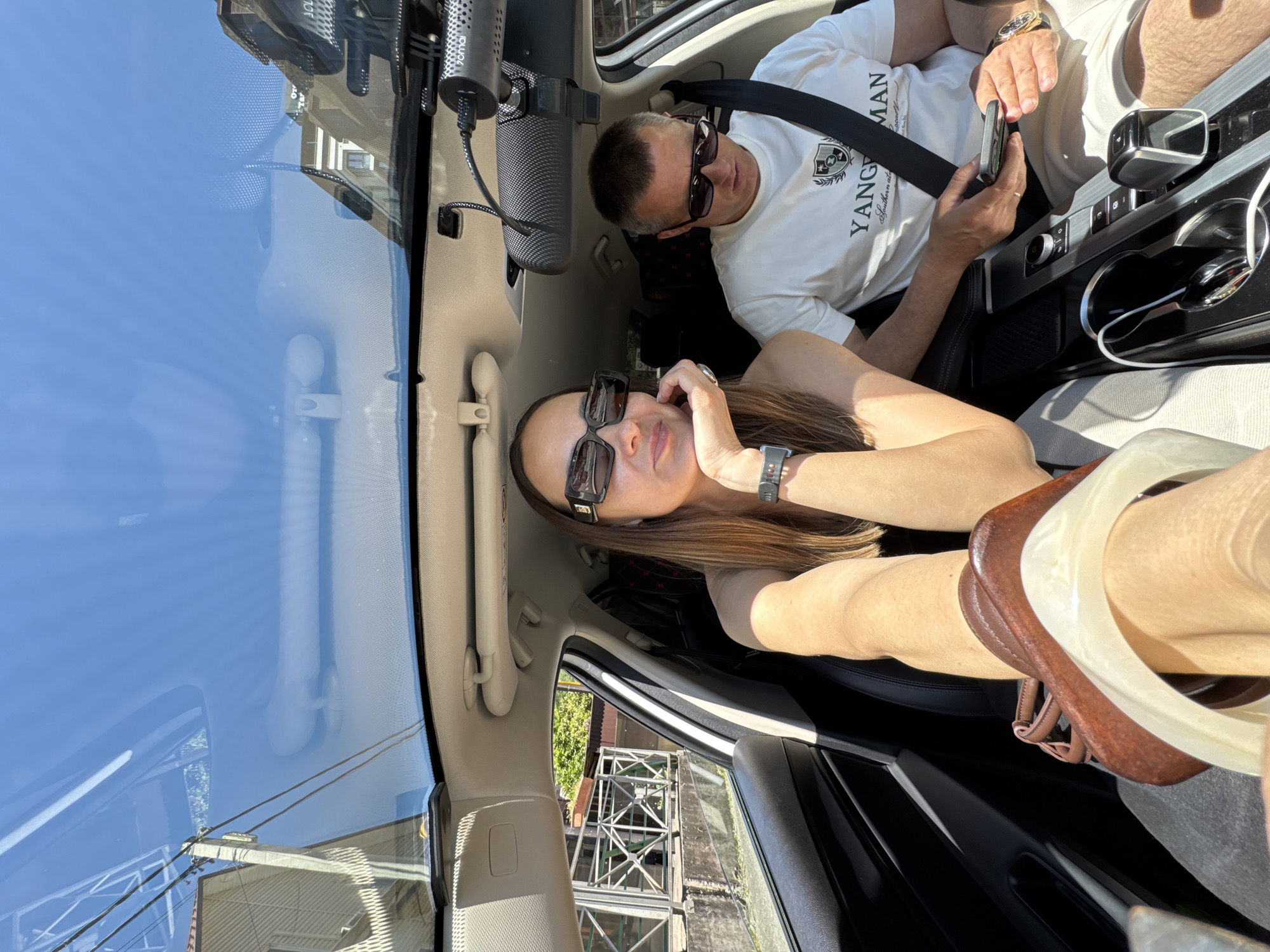 Two individuals, a woman and a man, are seated in the front of a modern car with a panoramic sunroof on a sunny day in the city of Sochi, Russia. The woman, positioned on the driver's side, has long brown hair and wears dark rectangular sunglasses, a dark top, a watch on her left wrist, and multiple bangles on her right arm. She rests her left hand on her chin, looking towards the camera. The man, on the passenger side, has short hair, wears dark sunglasses, a white T-shirt with a crest logo and "YANGMAN" text, and light-colored shorts. He is secured by a seatbelt and is looking down at a smartphone held in his right hand.
The car's interior features a central console with controls, cup holders, and a gear selector. A USB cable is connected. A "70mai" dashcam is mounted near the rearview mirror. Through the panoramic sunroof and windshield, reflections of a clear blue sky, utility poles, and power lines are visible. The background seen through the windows and reflections includes various multi-story buildings with different architectural styles (some with light siding, others with darker roofs and balconies), as well as green foliage and a metal fence structure, indicative of an urban or residential environment.