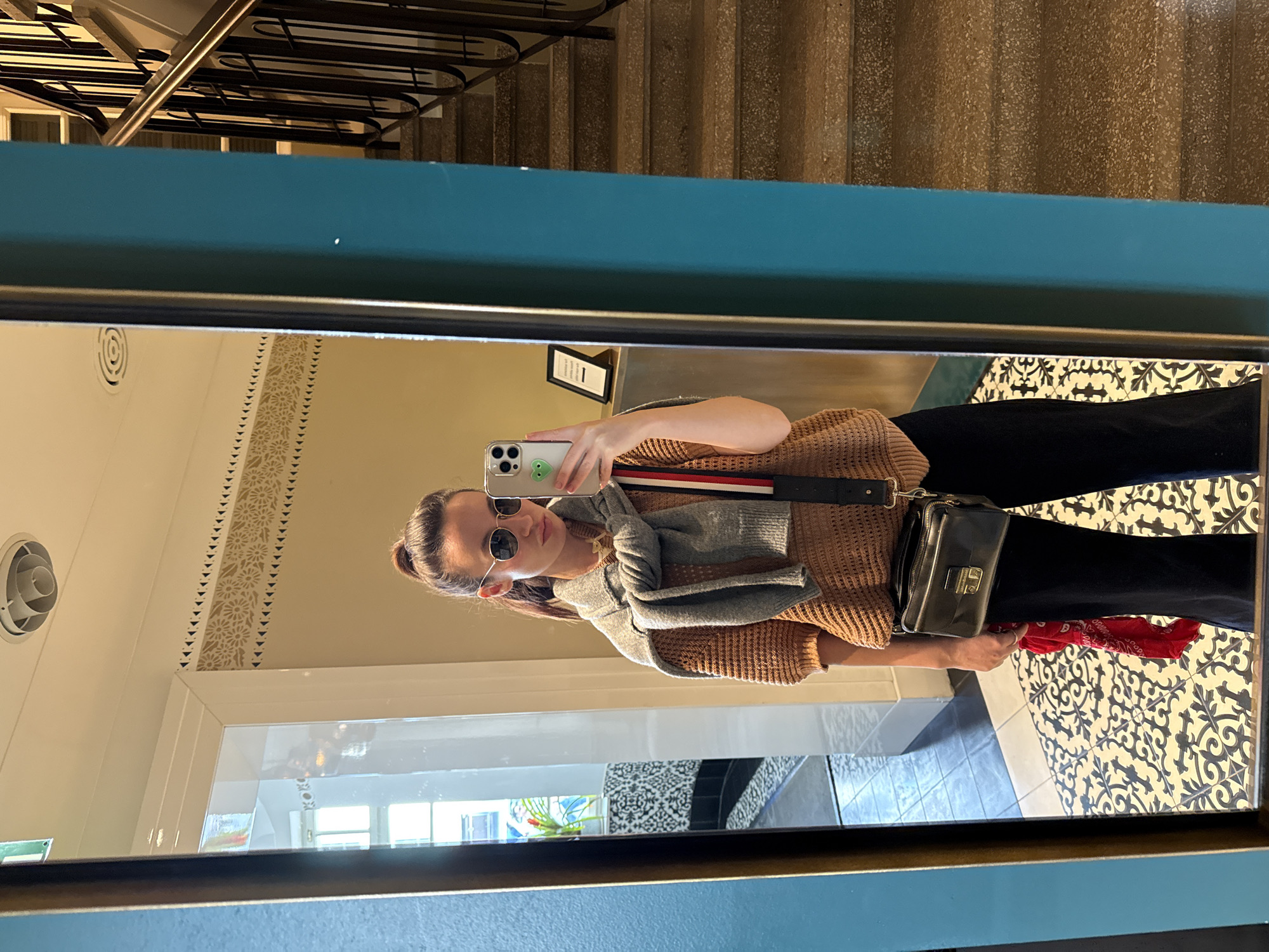 A woman is taking a mirror selfie in a hallway. The woman is wearing black pants, a brown sweater, and sunglasses, and a gray scarf. She has a black purse hanging from her shoulder. She's holding an iPhone in her left hand, and a red bandanna in her right. The hallway has a patterned tile floor, a white wall with decorative trim, and stairs are visible in the mirror's reflection. A sign is visible above her head, also in the mirror's reflection. While the location is not determinable based on the image alone, the user has suggested that the image was taken in Tel-Aviv, Israel.