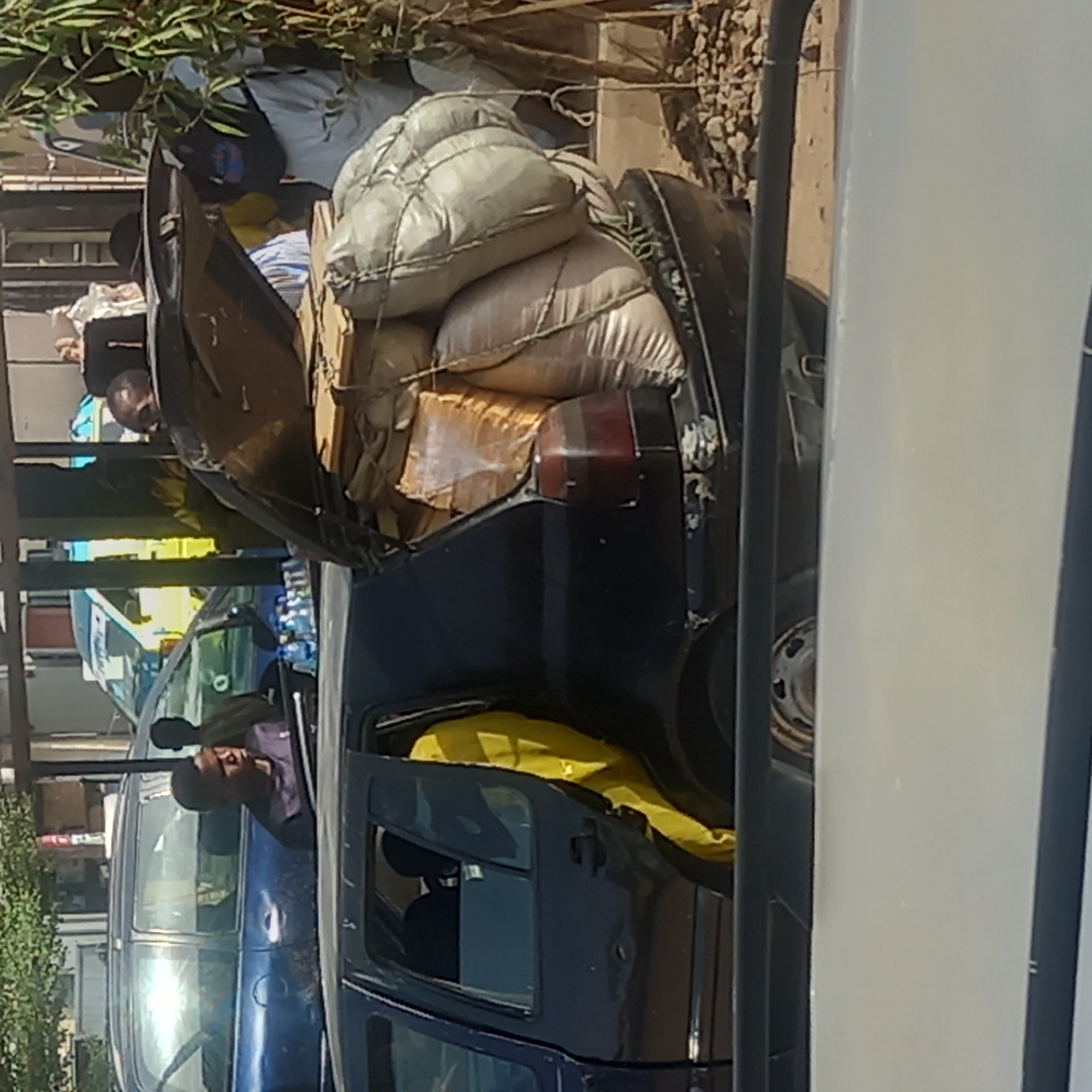 In Dadi Riba, Nigeria, a dark blue hatchback is overloaded with multiple large, white sacks secured with rope in the open trunk. The driver, a Black man, leans out of the driver's side window. A yellow object is visible inside the car. Other vehicles and buildings are in the background.