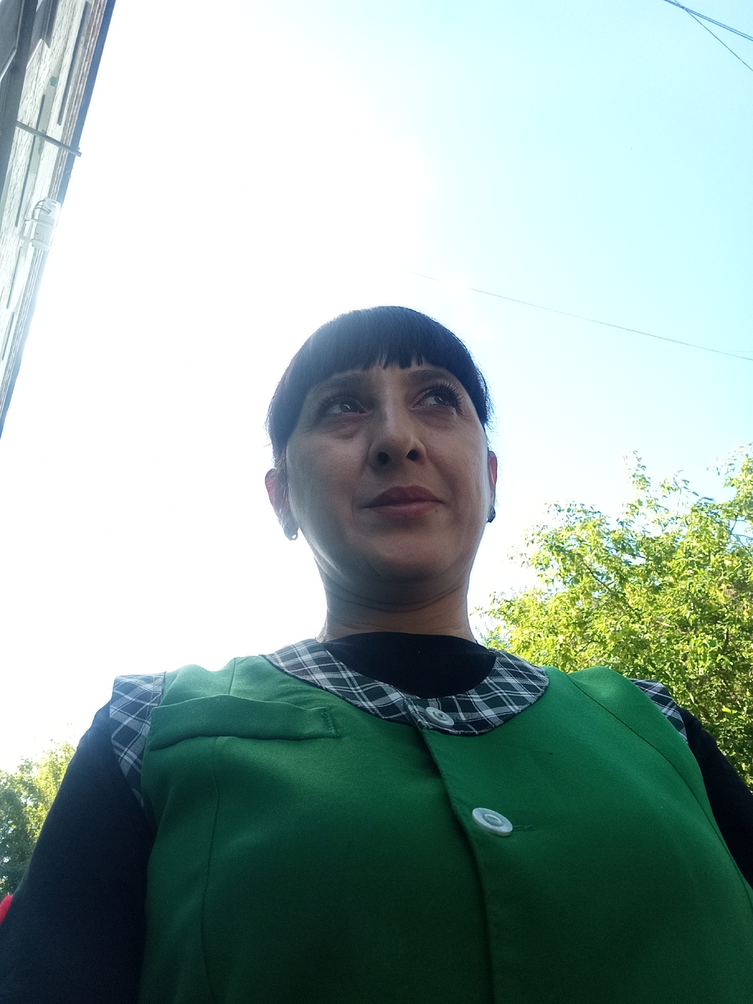 A low-angle, medium shot captures a woman from the chest up, looking upwards. She has dark hair with bangs and is wearing a green vest over a black shirt with a plaid collar. The background consists of a bright sky, a building on the left, and lush green trees on the right. The lighting suggests it is daytime, possibly with the sun behind the viewer. The image appears to be a selfie taken outdoors. It is not possible to determine the location in Belogorsk, Russia from the image alone.