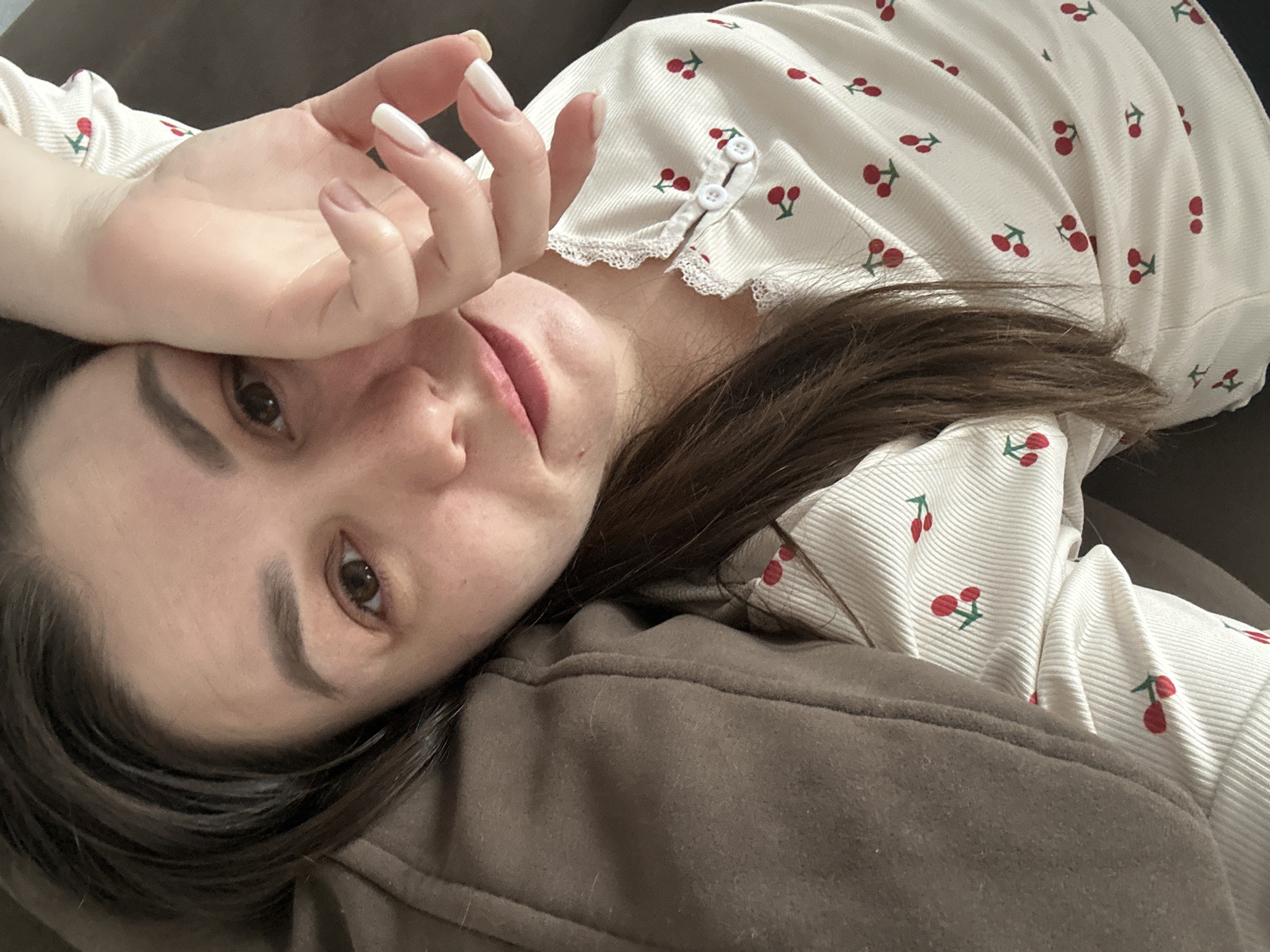 A woman is lying down, possibly on a couch or bed. She is wearing a white top with a cherry print and white buttons. Her left hand is raised near her face, obscuring part of it. The woman has dark hair and light skin. The background is brown and indistinct. The location and specific details about the city of Chaikovsky, Russia are not discernible from this image.