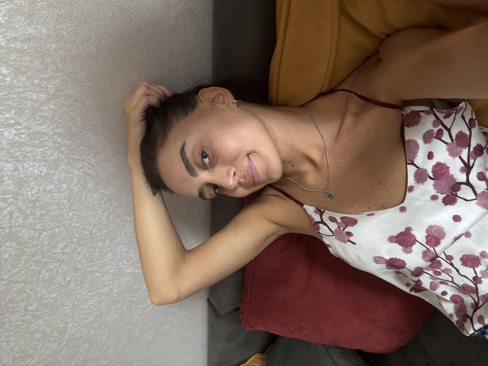 A woman with dark hair pulled back is lying on a couch with her arm raised behind her head. She is wearing a white camisole with a floral pattern and a silver necklace. The couch has a dark grey fabric, and there are two large pillows behind her, one red and one yellow. The wall behind the couch is textured and light grey. The scene appears to be indoors, likely in a residential setting. There is no information to suggest this location is in Tbilisi, Georgia.