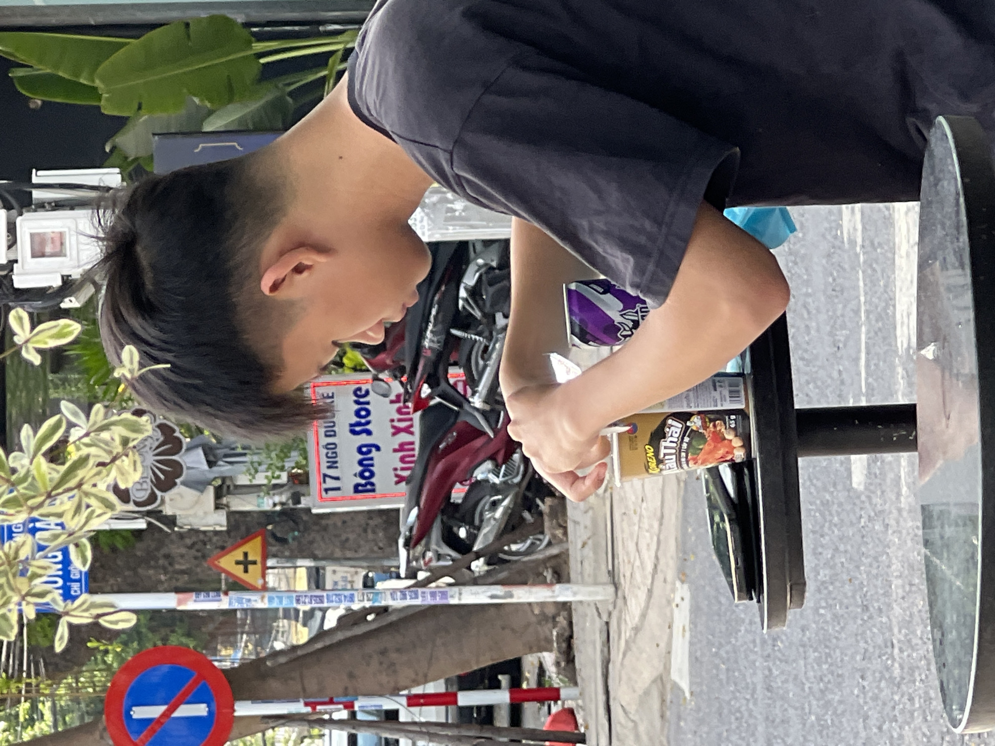 In Nha Trang, Vietnam, a young person with dark hair, styled with an undercut, sits at an outdoor table, focused on a cup in their hands. They wear a dark t-shirt. On the table, a cell phone and a cup labeled "KORENO 1ẩu Thái" are visible. Next to the cup, a can with purple and white branding is present. In the background, motorcycles are parked, and various signs are displayed on poles, including a "No Parking" sign, a yellow triangular sign with a cross, and a rectangular sign that reads "17 NGO DU KE Bong Store Vĩnh Xinh". Green foliage and large leaves are also part of the environment, suggesting a street-side cafe or shop.