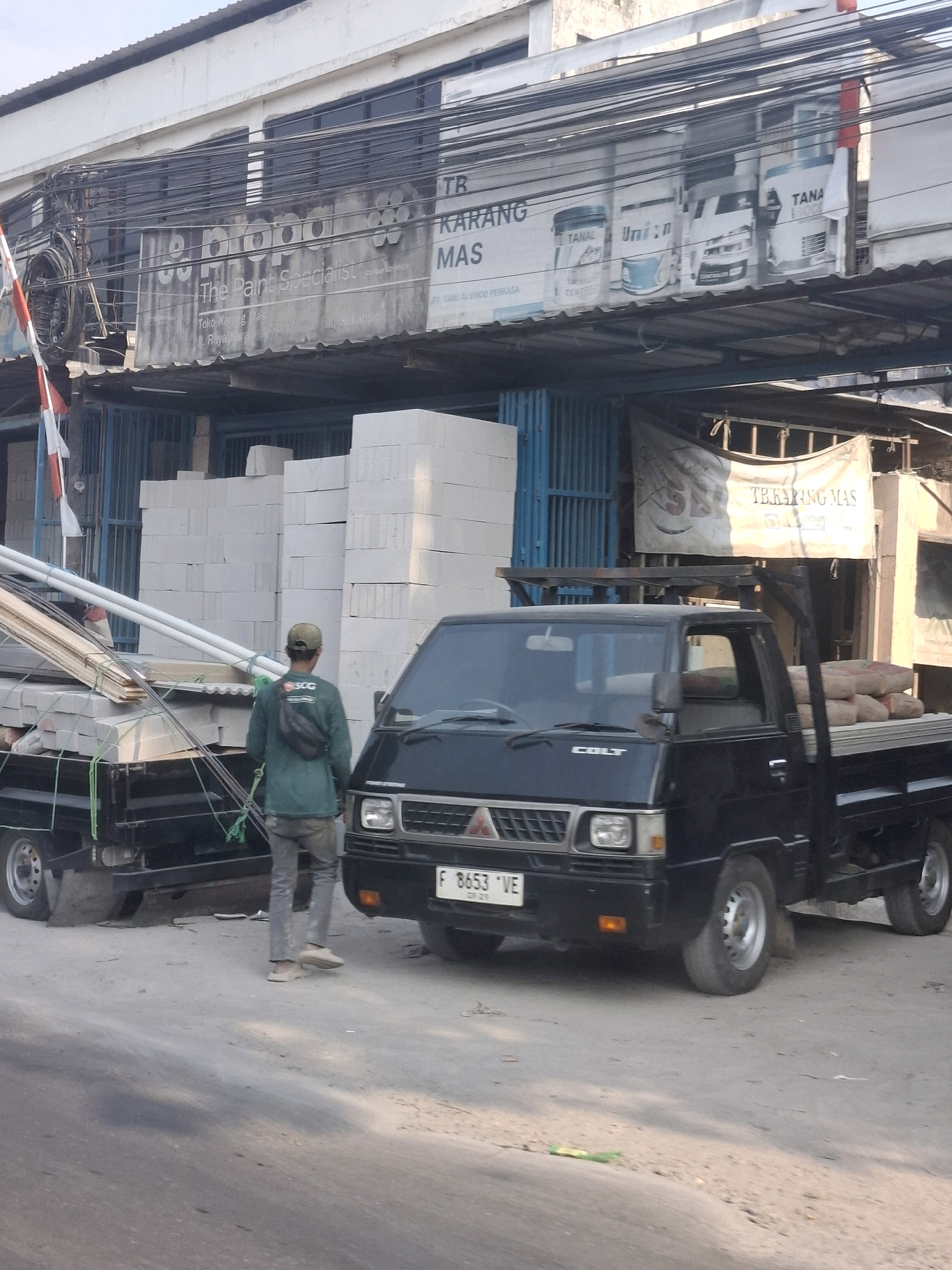 A flatbed truck, identified by the "COLT" logo on its grille, is parked on a dirt road in Karangtengah, Indonesia. The truck carries a load of building materials, including what appear to be bags of cement and stacks of white rectangular blocks. A man wearing a green long-sleeved shirt and gray pants is standing next to the truck, facing away from the camera. He has a black fanny pack around his waist. In the background, a building with various signs is visible, one of which reads "The Paint Specialist." Stacks of white concrete blocks are also piled near the building. A red and white striped flag is partially visible on the left side of the image. The scene appears to be an outdoor commercial area, possibly a building supply store or a construction site.