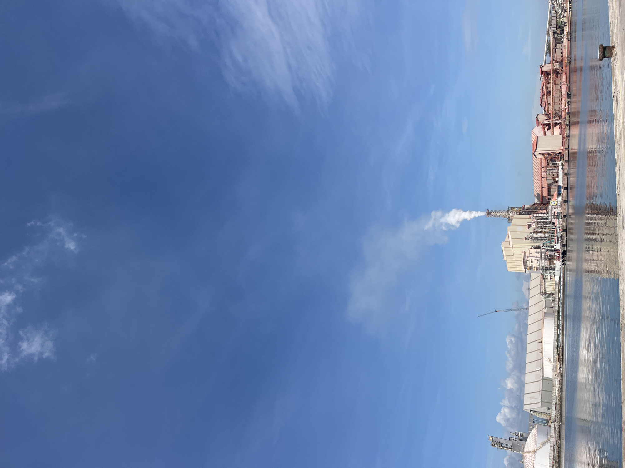 The image depicts an industrial landscape with a clear, blue sky filled with sparse, wispy clouds. A large industrial complex occupies the right side of the frame, reflecting in the body of water that borders it. A prominent smokestack rises from the complex, emitting white smoke. Various industrial structures, including buildings, tanks, and pipes, are visible within the complex. A crane is also present in the complex. The image is captured from a low angle. The industrial complex is located in Tambonbaroh, Indonesia.
