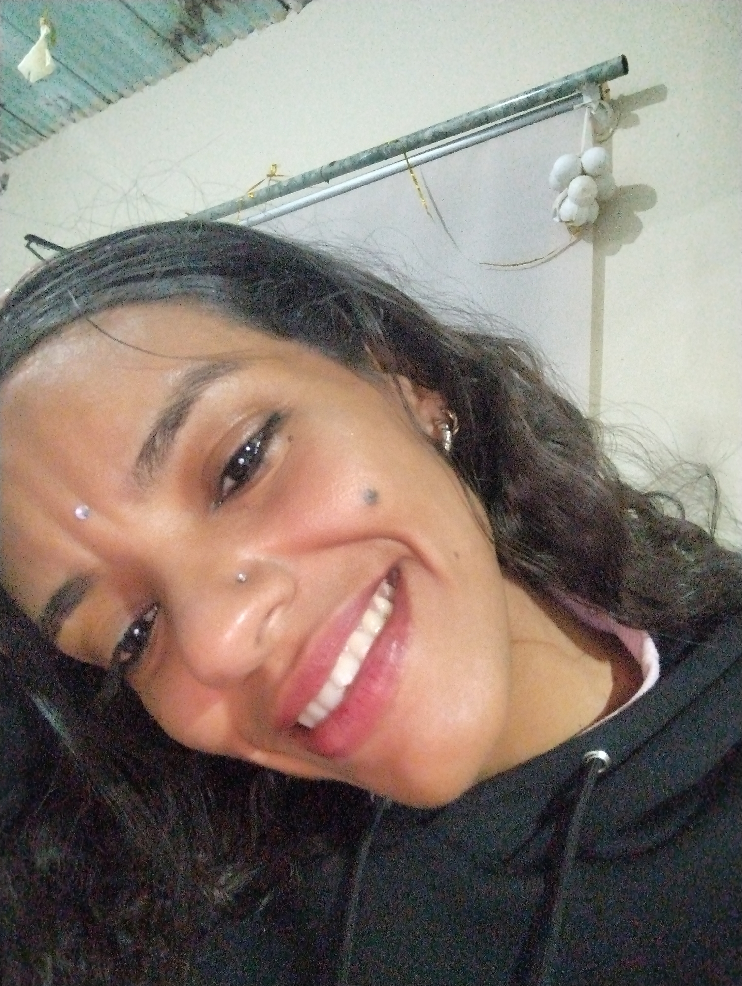 A close-up, low-angle shot of a woman with dark curly hair, fair skin, and a bright smile. She has piercings on her nose and forehead, a mole on her right cheek, and is wearing earrings. She is wearing a black hooded sweatshirt. The background includes a wall, a metal bar, and a bundle of white objects hanging from the bar. The location is El Valle del Espíritu Santo, Venezuela. The scene is indoors.