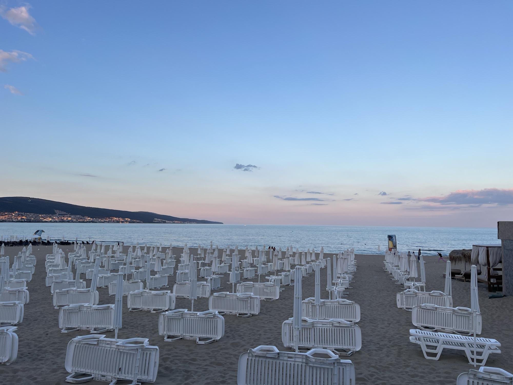 This image captures a beach in Nesebar, Bulgaria, during what appears to be early morning or late evening, indicated by the soft, diffused light and the colors in the sky. The beach is covered with rows of neatly stacked white plastic lounge chairs and white portable barriers, suggesting the area is prepared for or has recently concluded a busy period. Several closed white umbrellas are also visible among the chairs.
In the background, the sea is calm and stretches towards the horizon. A distant coastline with a city or town nestled on a hill is illuminated by soft lights, creating a warm glow against the darker silhouette of the land. The sky transitions from a pale blue at the top to soft pinks and oranges near the horizon, with a few wispy clouds scattered across it.
A few individuals are visible on the beach and near the water's edge, some walking and others seated at a distance. The overall scene is serene and orderly, with the natural beauty of the coast juxtaposed with the man-made amenities of the beach. The perspective is from the sand, looking out towards the sea.