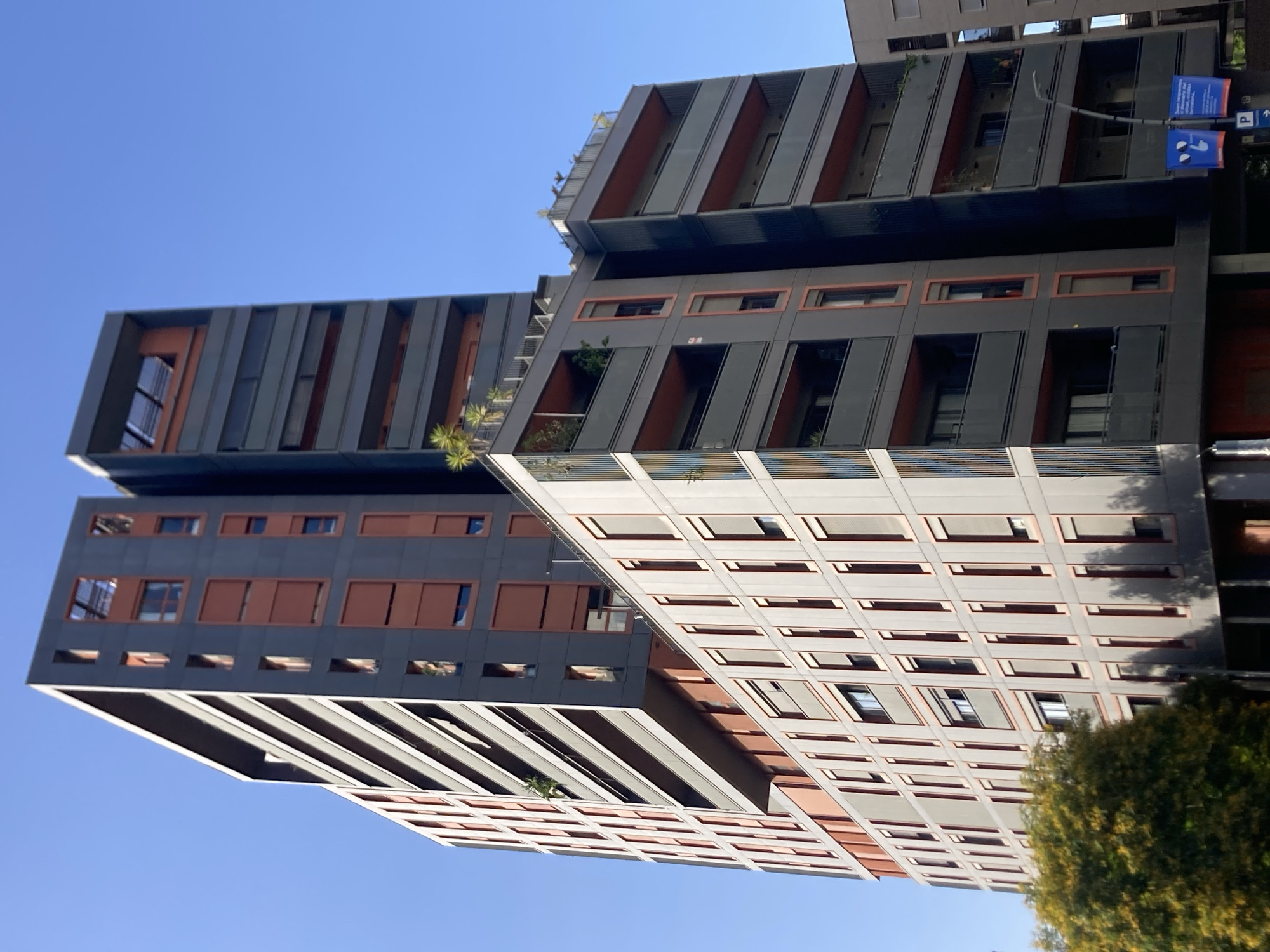 This media file captures a bright, sunny day in Barcelona, Spain, with a clear blue sky dominating the background. The scene focuses on a modern architectural complex of multi-story buildings. The buildings exhibit a striking design with alternating sections of dark grey panels and reddish-orange frames around windows and balconies. Some sections feature vertical slats, adding to the contemporary aesthetic.
Several balconies are visible, some adorned with greenery, suggesting residential or commercial spaces. The angles of the buildings create a dynamic visual impression, with one structure appearing to lean or jut outwards. On one of the buildings, there are two blue signs, one with an image of a person and another depicting a parking symbol. The parking sign also has text, but it is not clearly legible in the image. A large, lush green tree with yellow-orange foliage is visible in the lower right corner, providing a contrast to the urban structures. The orientation of the image is such that the buildings appear to be viewed from below, looking upwards towards the sky. The sunlight suggests it is daytime, likely late morning or early afternoon. No people are clearly visible in the frame, and the overall impression is one of modern urban living bathed in pleasant weather.