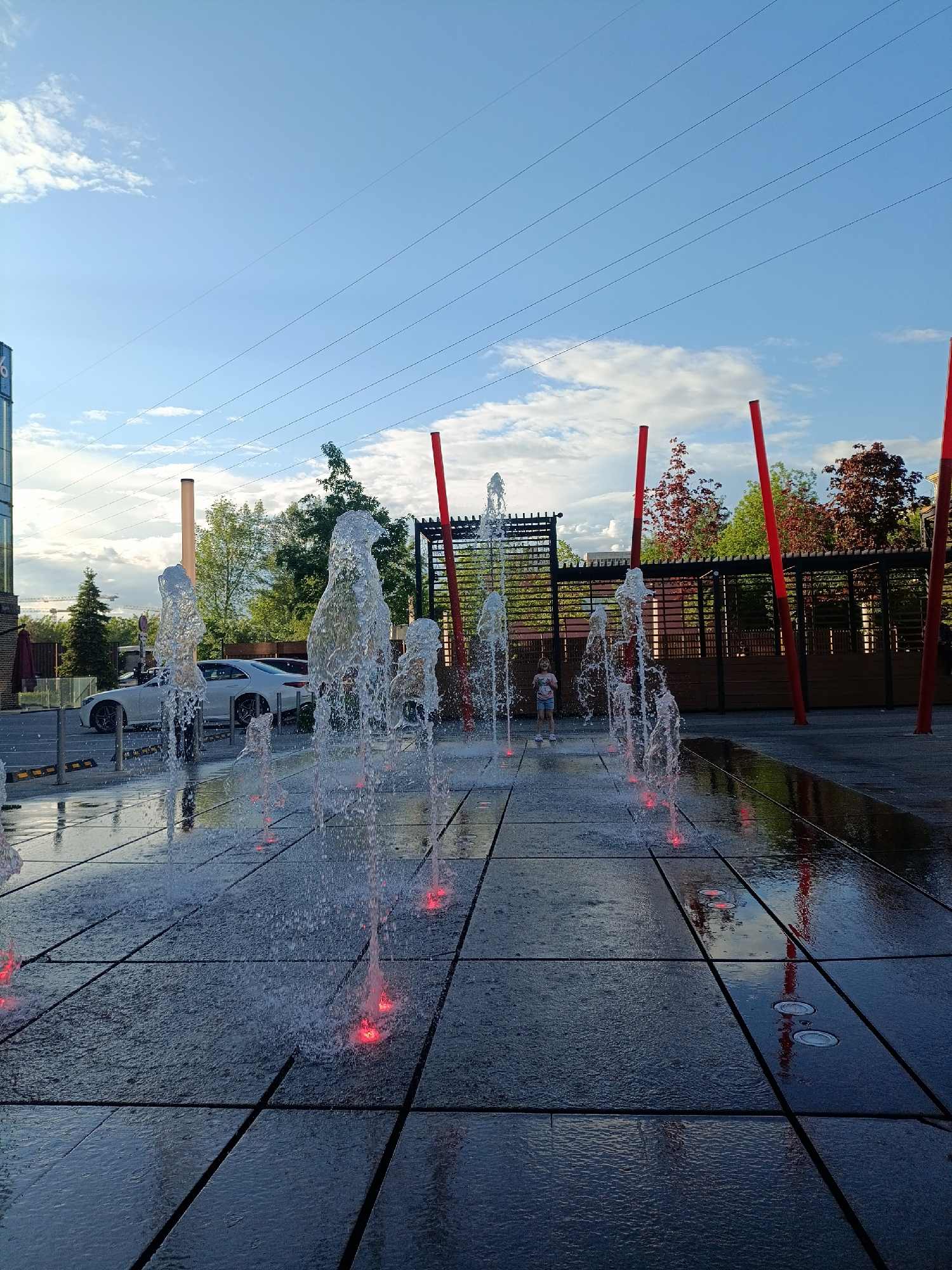 The image shows an outdoor water fountain area in what appears to be a public space. Water jets are active, with some illuminated by red lights at the base. A child is standing near the fountain, seemingly interacting with the water. The ground is paved with large, dark tiles reflecting the sky and surrounding structures. In the background are trees, architectural elements including a structure with red pillars and a trellis, and a white car parked along the perimeter. Overhead power lines are visible against the blue sky with scattered clouds. The image depicts a lively, urban scene on a clear day. Based on the context, it is likely to be located in Moscow, Russia.