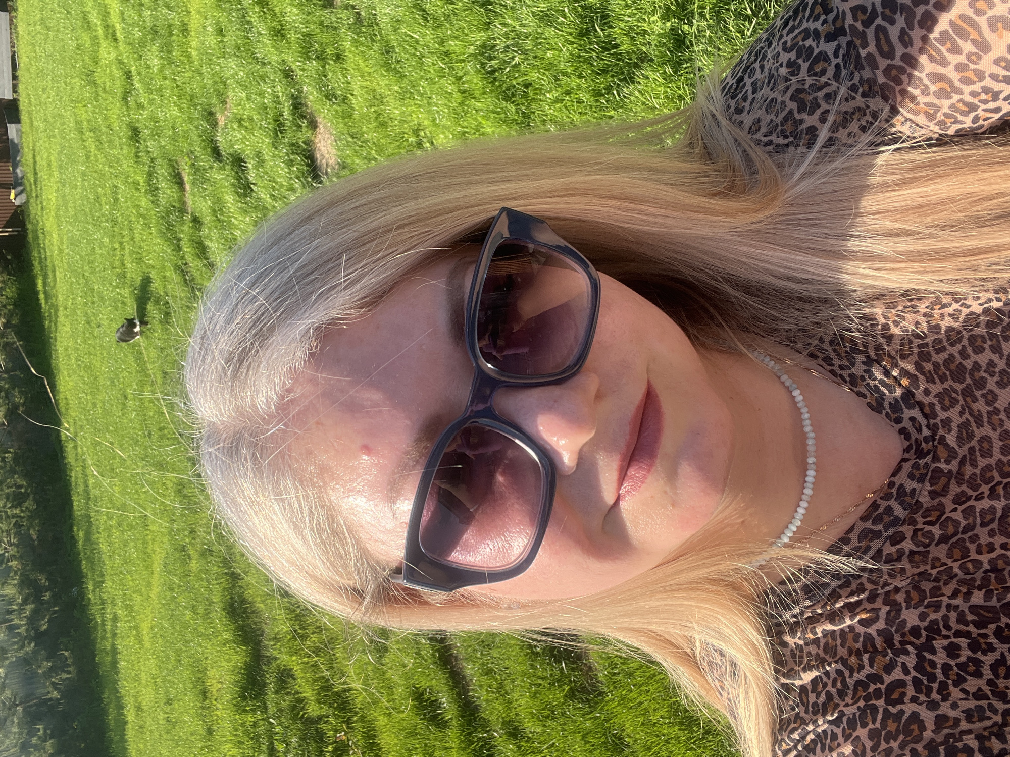 The image is a close-up, eye-level selfie of a fair-skinned woman with blonde hair wearing sunglasses and a leopard-print top. She is wearing a white beaded necklace, possibly a choker. The background shows a patch of green grass, suggesting she is outdoors. There's a small dark object, possibly a bird or animal, visible in the grass. The lighting indicates it is a sunny day. The location is not evident in the image, and there is no reference to Новая Чернушка, Russia.