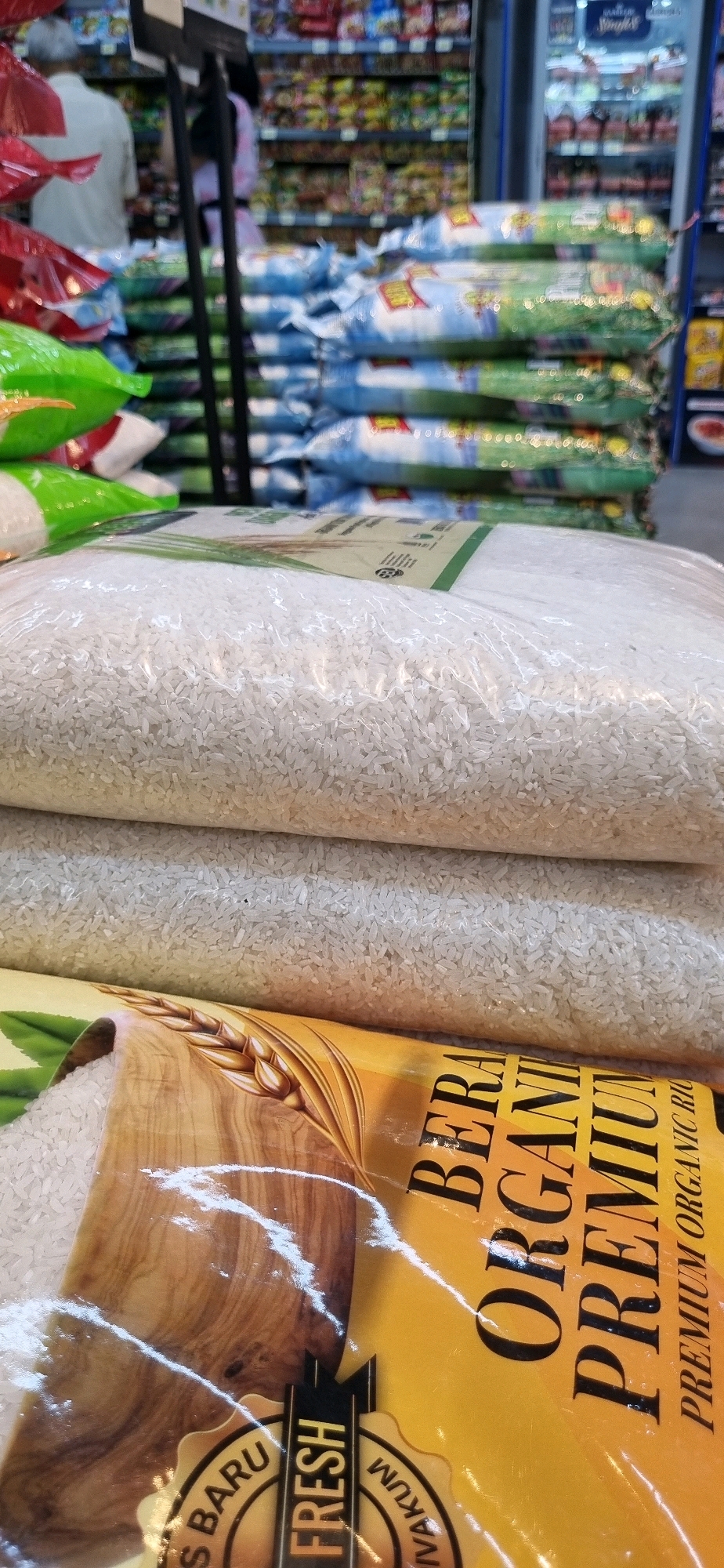 In a supermarket aisle in Denpasar, Indonesia, several large, clear plastic bags of white rice are stacked on shelves. The bags are partially filled, with the rice visible inside. The frontmost bag is a premium organic rice brand, indicated by text on the packaging that reads "BERAS ORGANIK PREMIUM" and "FRESH." In the background, other rice bags are visible, along with shelves stocked with various food items. Two individuals are partially visible in the background, suggesting ongoing shopping activity. The lighting is typical of a commercial establishment, with overhead lights illuminating the products.