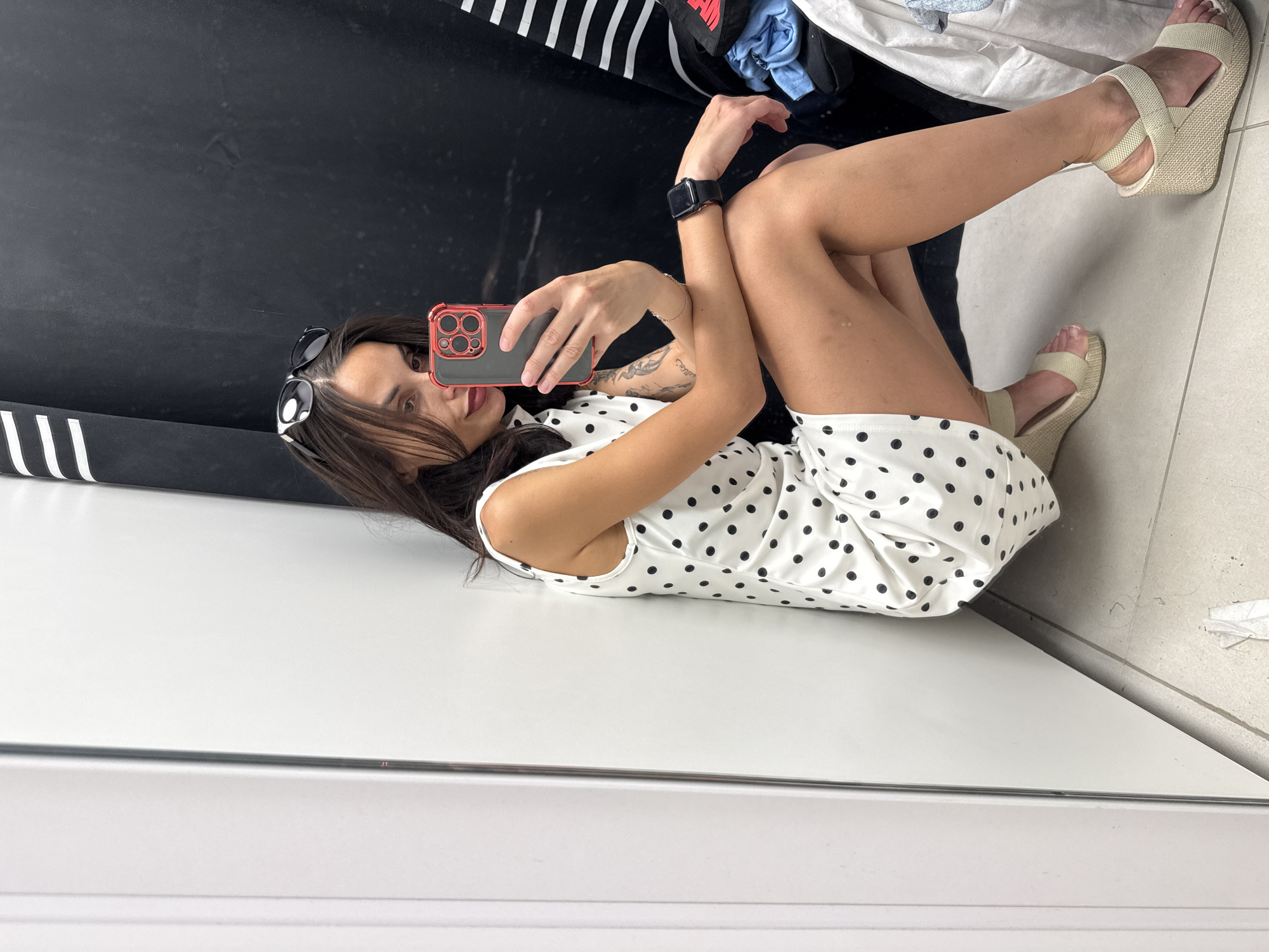 A woman is taking a selfie in a room. She is wearing a white dress with black polka dots and beige wedge sandals. The woman is holding a red and black smartphone and has sunglasses perched on her head. She has a black smartwatch on her left wrist and visible tattoos on her left arm. The room has a black and white striped garment rack and assorted clothes. The floor is tiled, and the walls are a mix of white and dark surfaces. The photograph may have been taken in Pleven, Bulgaria.