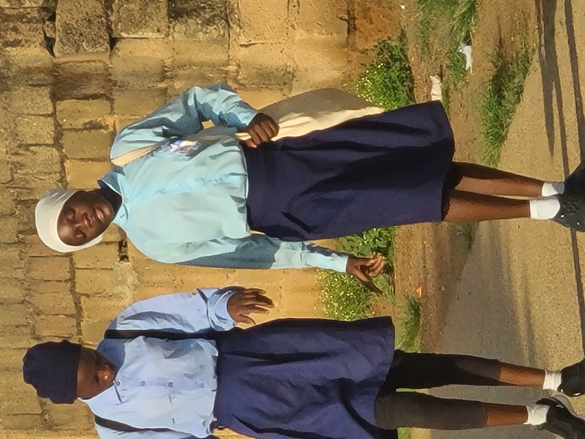 Two young girls, likely students, walk side-by-side in front of a rough-hewn stone wall. The girl on the left wears a dark blue beanie and a light blue collared shirt with dark blue suspenders and a dark blue skirt over black leggings. The girl on the right wears a white headscarf and a light blue collared shirt with a dark blue skirt. She carries a light-colored cloth bag over her right shoulder. Both girls wear white socks and dark shoes. The ground is a mix of dirt and paved surface. Green grass and small white flowers are visible in the background, suggesting a location with some vegetation, possibly a school or residential area in Jos, Nigeria. The lighting indicates it is daytime.