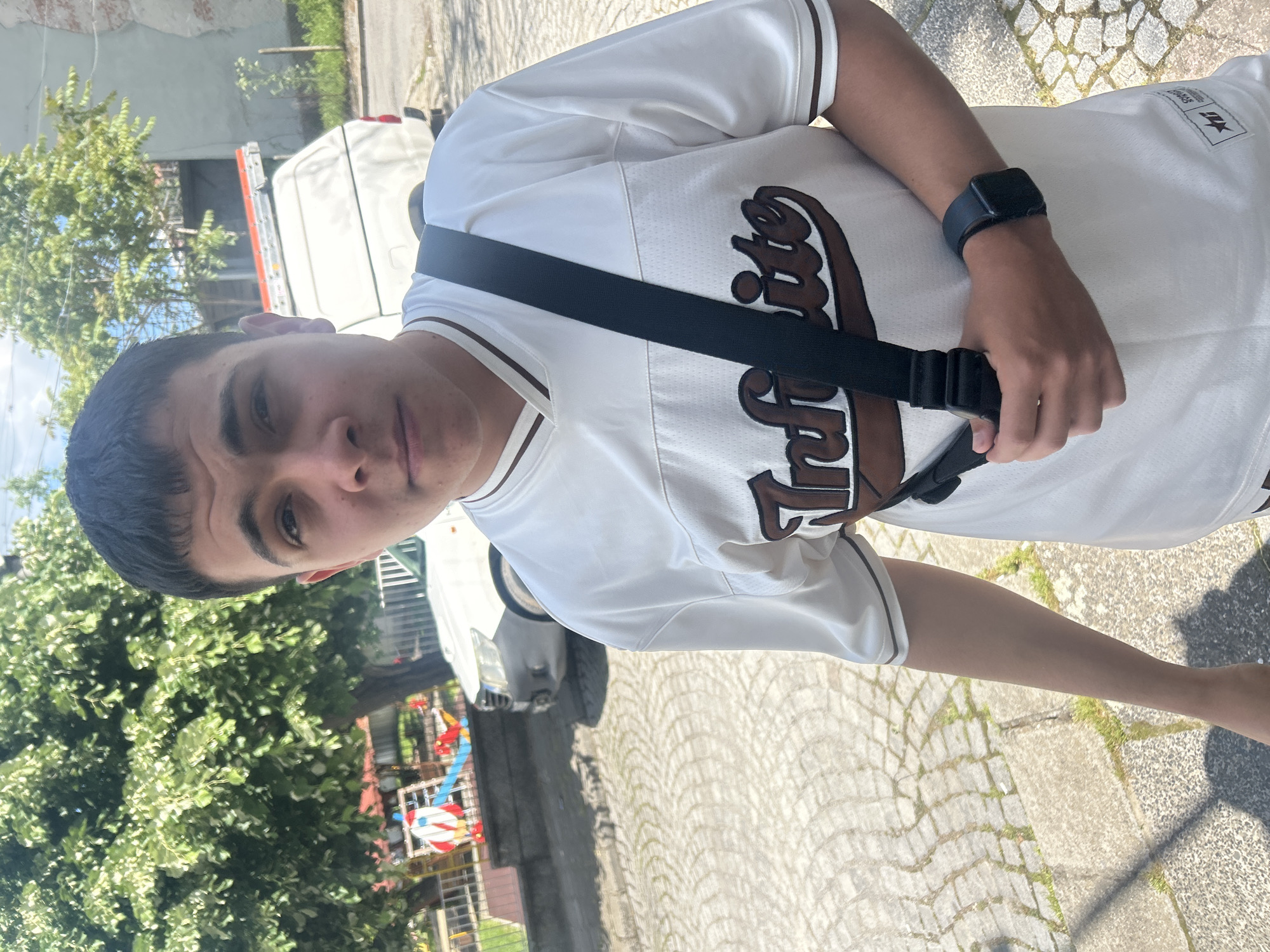 A young man is standing on a cobblestone sidewalk in Elhovo, Bulgaria. He is wearing a white baseball jersey with brown lettering and trim, a black crossbody bag, and a black smartwatch. A white van and a silver car are parked on the street behind him. In the background, there is a green tree, a brick building with peeling paint, and a colorful play structure.