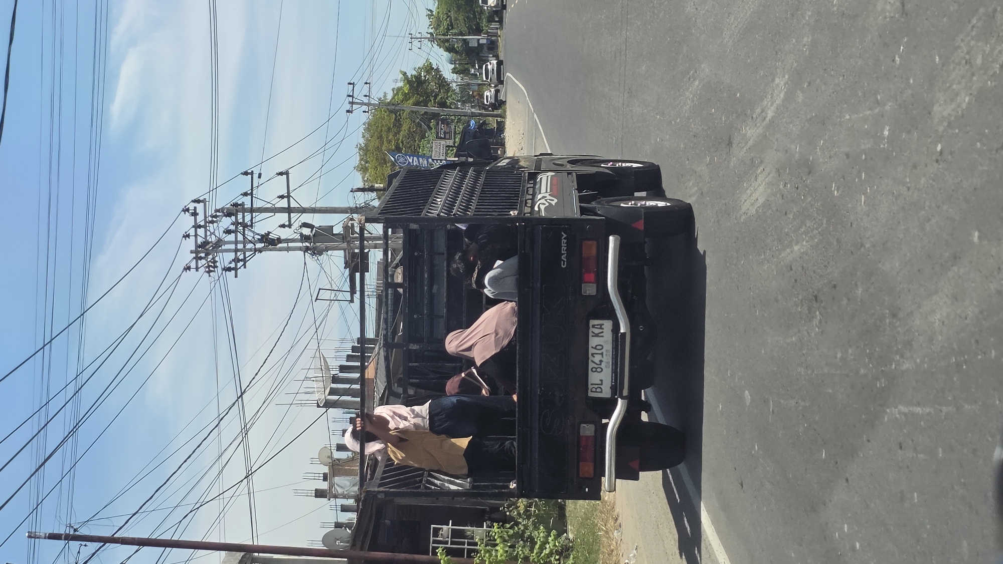 The image depicts a black "Carry" pickup truck moving on a paved road. Several people are riding in the back of the truck, which has a metal cage structure. A man in a light-colored shirt and white head covering is standing near the back of the truck, while others are seated. The license plate reads "BL 8416 KA." Overhead, there are power lines and utility poles. In the background, trees and other vehicles are visible. The location is Cotgadong, Indonesia.