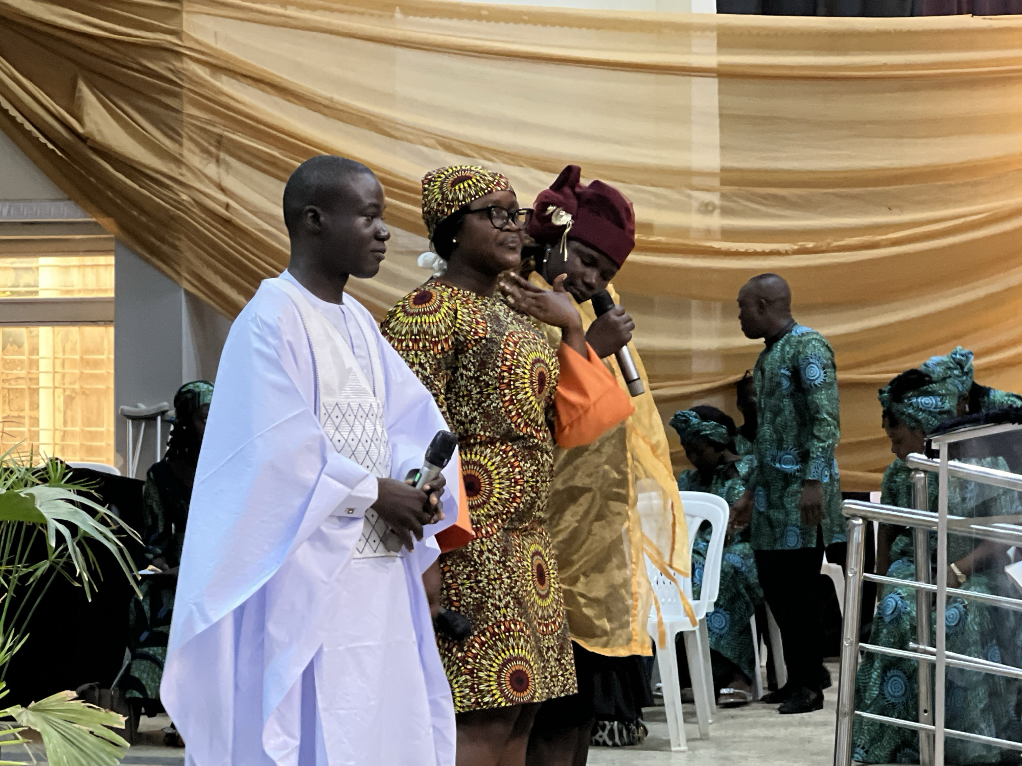 The image captures an indoor event with a backdrop of draped golden fabric. In the foreground, four people are visible, three of whom are standing and facing left. A man on the far left wears a white traditional garment and holds a microphone. Next to him, a woman in a patterned dress and headscarf is speaking, also holding a microphone. To her right, another individual in a maroon head covering and a golden garment is seen from the side. Further to the right, a man in a green and blue patterned outfit stands, facing left. Several other individuals in similar green and blue patterned attire are seated or standing in the background, along with white plastic chairs. The lighting suggests an artificial light source. The image appears to be taken in Kabong, Nigeria, possibly at a community gathering or ceremony.