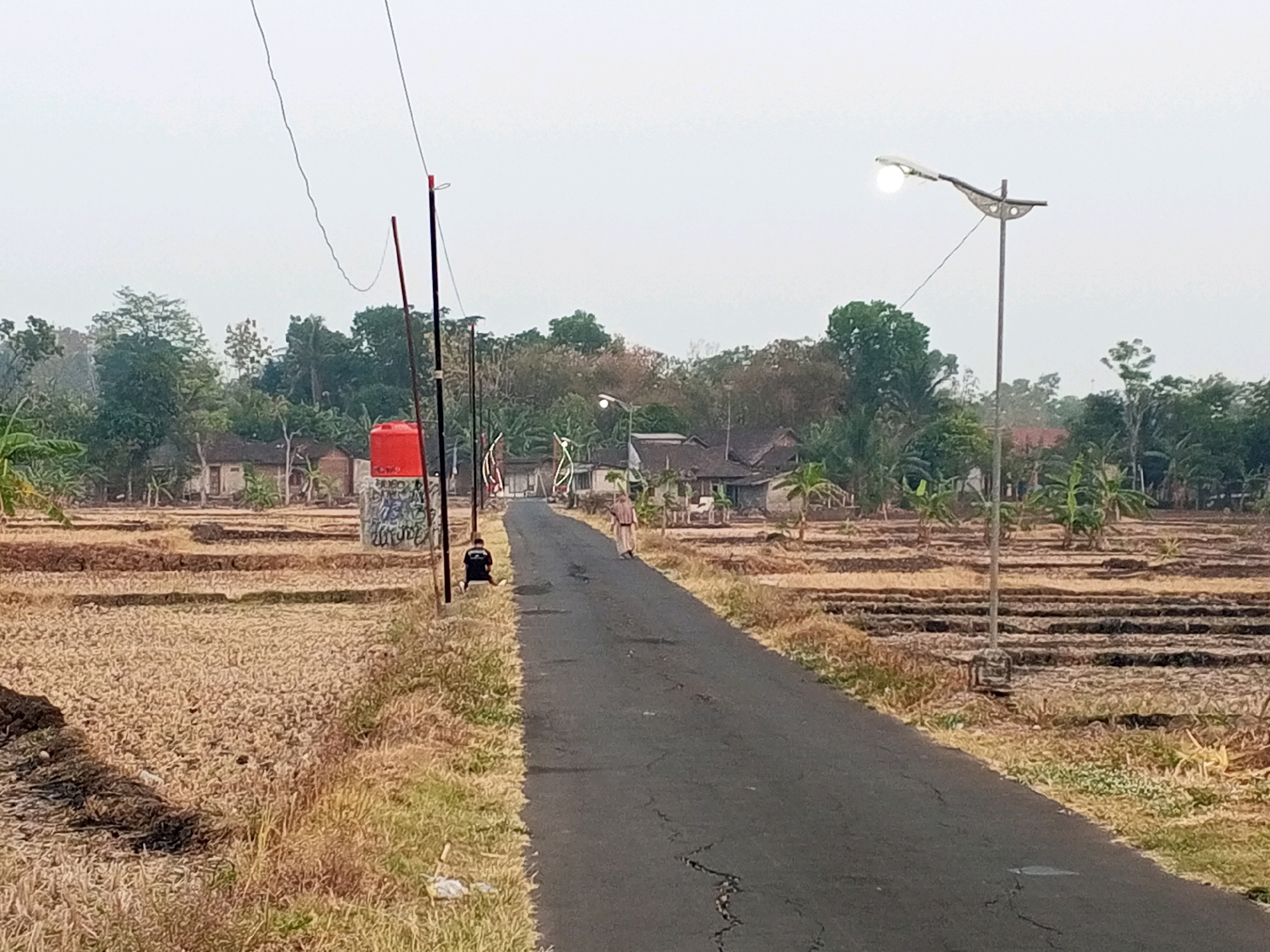 This image depicts a rural road in Klari, Indonesia, likely during the daytime given the light. The road is paved and appears somewhat worn, with visible cracks. To the left of the road, there are dry, tilled fields, possibly recently harvested or prepared for planting, with scattered dry vegetation. A tall, red water tank is situated to the left, near a pole with graffiti on it. A person in dark clothing is seated on the ground next to the pole. Further down the road, a person in a light-colored, loose-fitting garment is walking away from the camera. The right side of the road also features tilled fields and some vegetation. In the background, there are several houses with traditional Indonesian architecture, surrounded by trees and banana plants. A modern street lamp with a lit bulb is visible on the right side of the road, taller than the surrounding structures. The sky is overcast and hazy. The overall impression is of a quiet, rural setting.