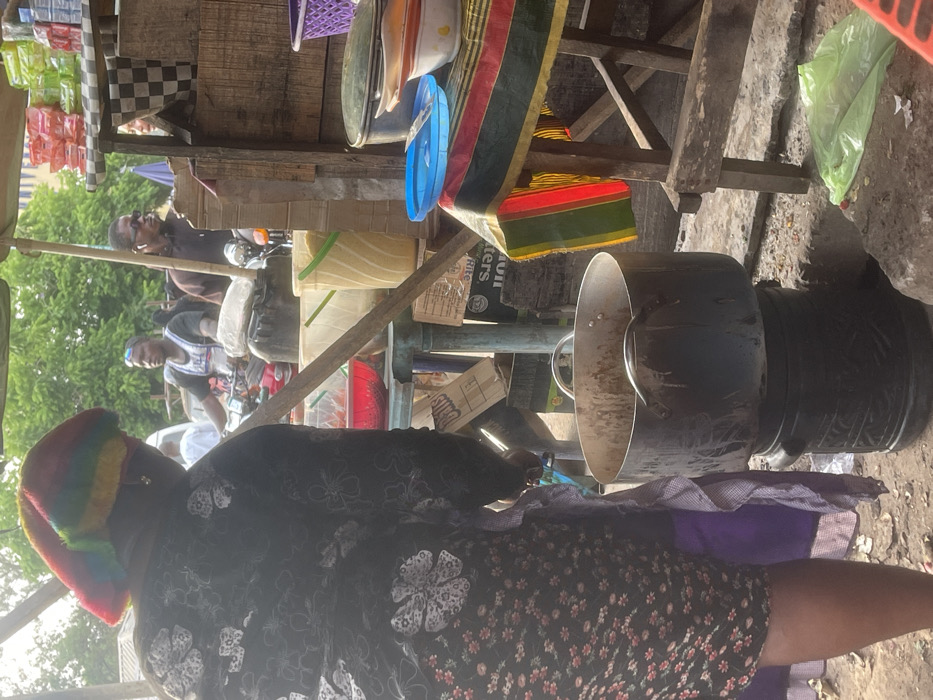 The image captures an outdoor market or street stall scene during daylight hours. The perspective is from behind a person in the immediate foreground, looking towards the stall and other individuals.
In the foreground, a person is seen from the back, wearing a dark-colored garment with a white floral or abstract pattern. They have a distinctive, colorful hat or head covering with horizontal stripes in hues of red, orange, yellow, green, and blue/purple. A purple fabric is visible to their right (viewer's left), possibly part of their attire or an item they are carrying. The ground appears to be a mix of dirt and concrete.
To the right of the foreground person, a prominent black, cylindrical machine or large pot stands on the ground. It has a metallic (silver/grey) funnel-like top or lid and a handle. This object suggests some form of food preparation or processing is taking place at the stall.
The midground features a rustic wooden stall structure with shelves and support beams. Various items are displayed or stored on and around it. On the upper parts of the stall, there are purple plastic baskets, white or cream-colored bowls, and a blue plastic lid. A piece of striped fabric, possibly depicting Pan-African colors (red, yellow, green, and black) or a Kente-style pattern, hangs from the stall. Below, other containers, including what appears to be a light yellow-green plastic bucket and a red container, are visible.
Several people are in the midground and background, interacting with the stall or passing by. One man, wearing a dark shirt and sunglasses, is seated on what appears to be a motorbike. Behind him, another man in a white sleeveless top with a pattern, possibly a jersey, is also visible. Both are looking towards the right side of the frame. Green foliage from trees or bushes is visible in the background, reinforcing the outdoor setting.
Visible text is limited and partially obscured. On a cardboard box located underneath the stall, the word "BUTTERS" is partially legible, with "OIL" or "SOIL" possibly below it. Another box shows partial letters "D" and "S".
The scene conveys the bustling atmosphere of a local market or roadside vendor, characterized by makeshift structures, various goods, and everyday activity.