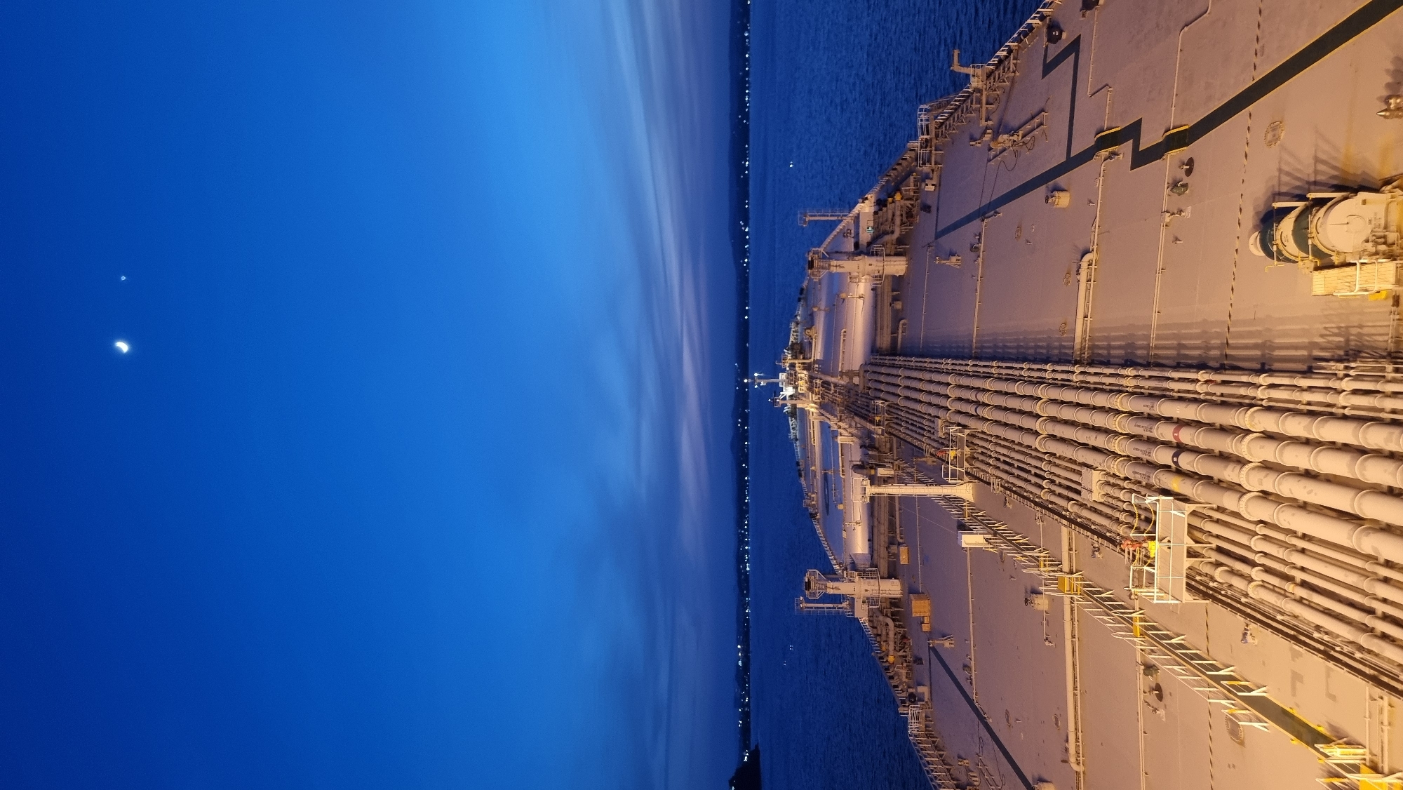 The image is a high-angle shot of the deck of a large cargo ship at dusk. The ship's deck is covered in a complex network of pipes, which are illuminated by artificial lights. A person wearing a yellow vest is visible on a platform among the pipes, seemingly performing maintenance. In the distance, the water stretches to the horizon, where a city's lights are visible along the coastline. The sky is a deep blue, with a crescent moon and a bright star or planet visible near the top of the frame. The overall scene suggests a working port environment at the transition from day to night.