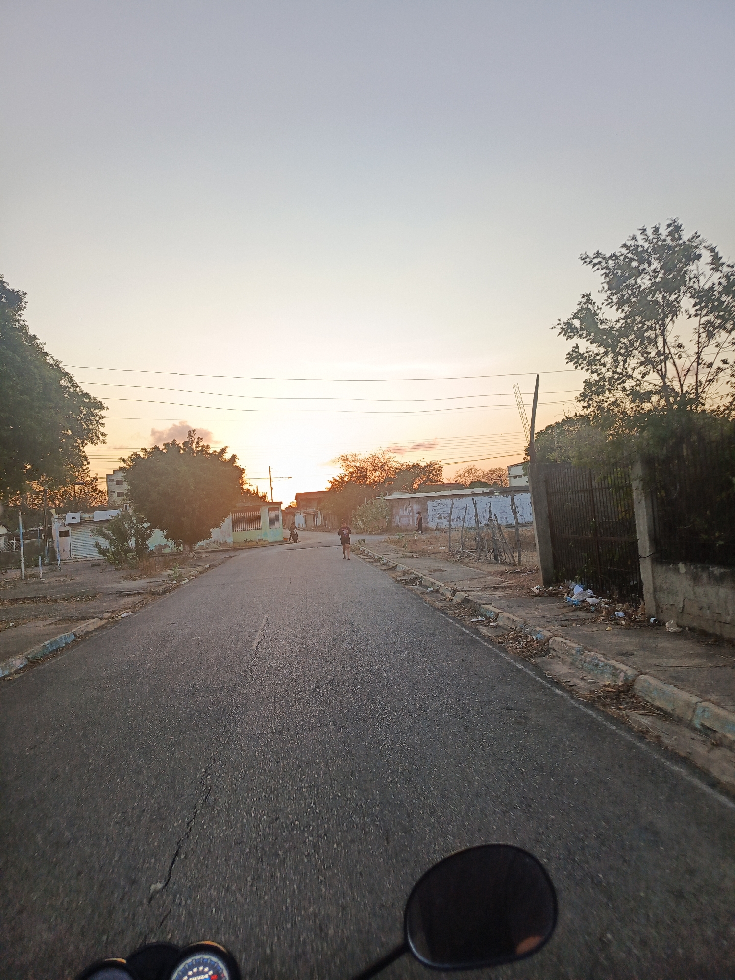 Captured from a low angle, presumably from a moving vehicle, the image presents a street view during a sunset in Juan Griego, Venezuela. A dark asphalt road dominates the foreground, exhibiting cracks and leading towards a visible sunset. On either side of the road, the curb and sidewalk are lined with debris and foliage. Buildings are set back from the road, appearing mostly rundown with aging facades. The sky is colored with a gradient of hues from the setting sun. A lone person is walking along the side of the road. Power lines are visible across the sky. The mirror of the vehicle taking the picture is visible in the foreground.