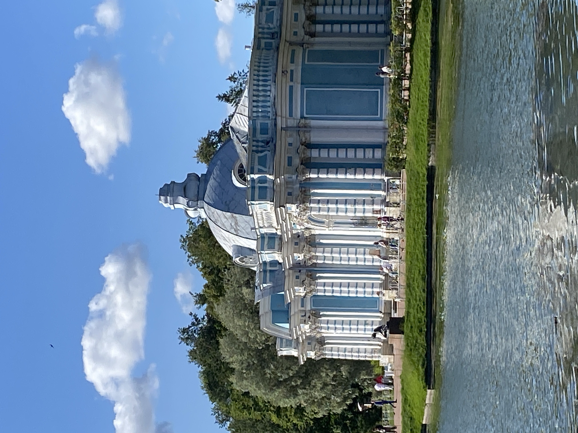 This is a full-shot, eye-level, outdoor photograph of the Hall of Apollo at the Catherine Palace in Pushkin, Russia. The architectural structure, painted in a striking light blue and white, is situated on the edge of a calm body of water, likely a pond or lake. The building features a prominent dome and a series of ornate columns and balconies. In the foreground, the water reflects the blue sky and white clouds. The background is filled with lush green trees. Several people are visible in the scene, some walking on a path beside the building, others sitting on benches or near the water. One person appears to be flying a small object in the sky, possibly a drone or a kite. The sky is a clear blue with fluffy white clouds.