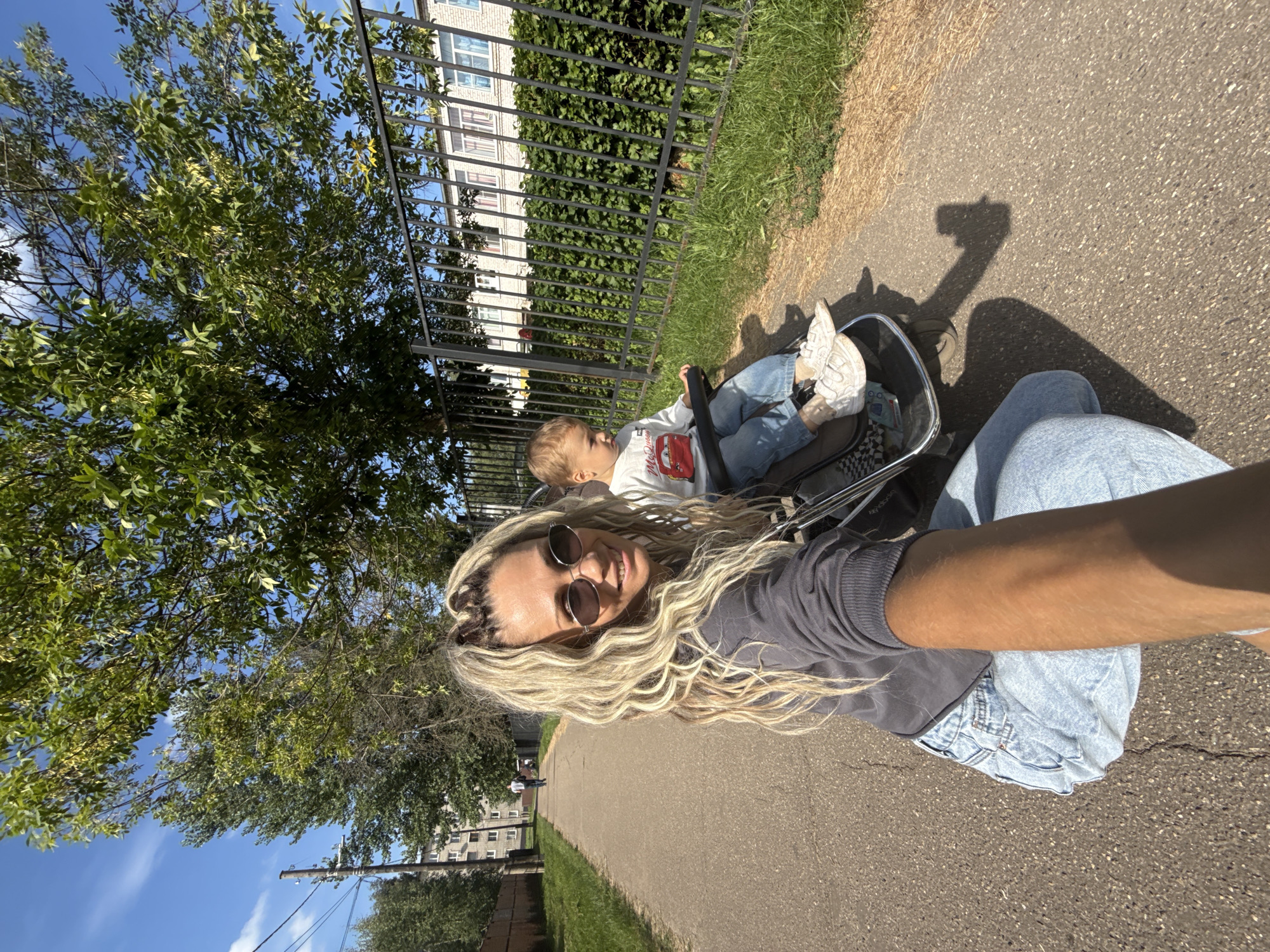 In the city of Rusanovo, Russia, on a sunny day, a woman with long blonde curly hair, wearing sunglasses and a grey sweatshirt, is taking a selfie while pushing a child in a stroller along a paved path. The child, dressed in a white long-sleeved shirt and blue jeans, is seated in the stroller. To the right of the path, a black metal fence borders a grassy area and a building with multiple windows. Trees with green leaves are visible behind the fence and in the background. The path leads towards buildings and trees, with a utility pole and wires also present in the distance. The woman's arm is extended, holding the camera.