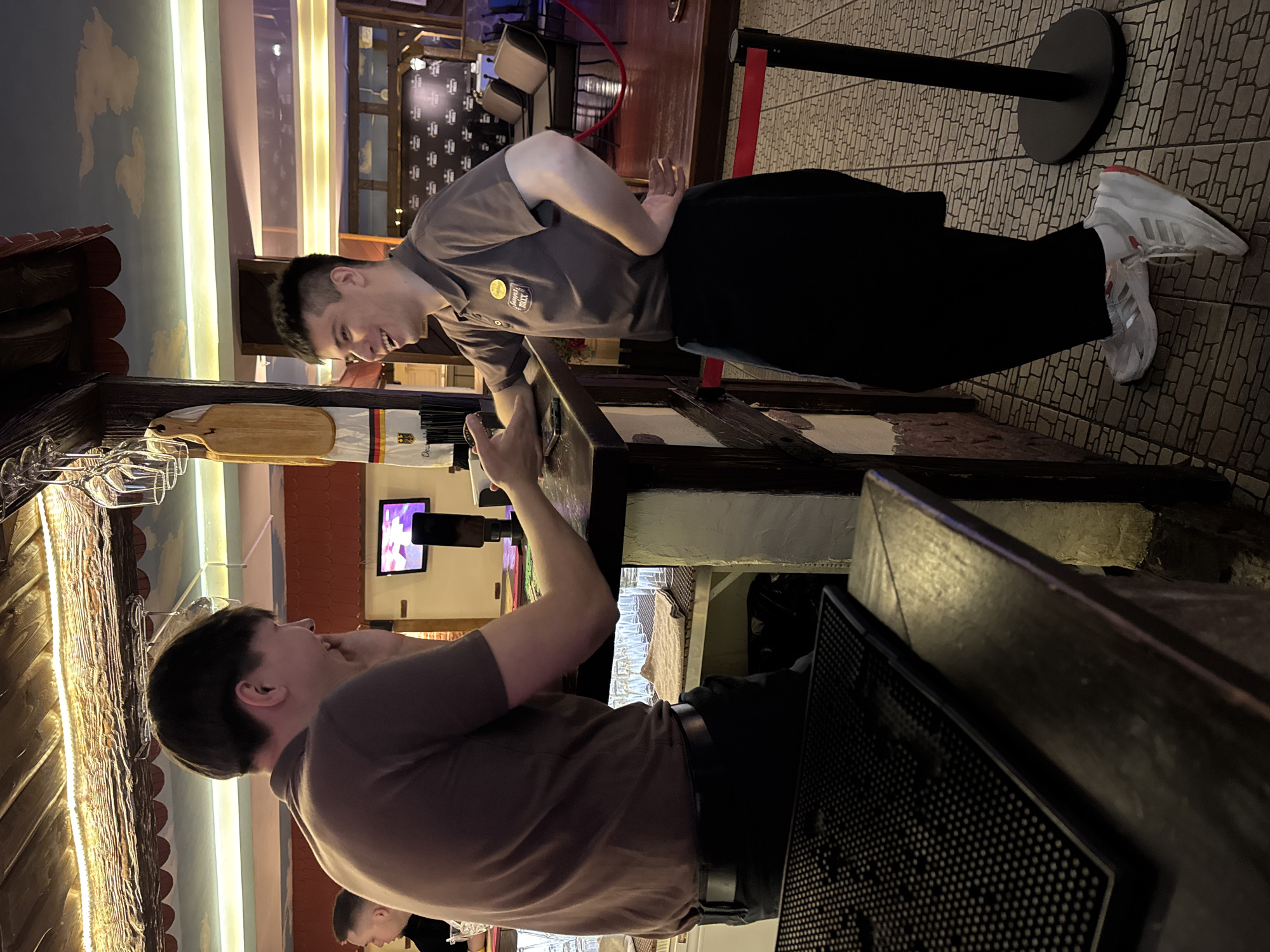 The image depicts an interior view of Samba Bar in Chaikovsky, Russia. Two adult males are engaged in conversation at a dark wooden bar counter. The man on the left, identified as a patron, wears a dark brown polo shirt and dark trousers. His right arm rests on the counter, with a dark smartphone nearby, and his left hand is near his chin as he smiles toward the man on the right. The man on the right, a staff member, wears a dark grey polo shirt with a yellow badge identifying "SAMBA BAR ЧАЙКОВСКИЙ" and a black apron. He stands behind the bar on a patterned tiled floor, wearing white Adidas athletic shoes with red accents. His left hand is placed on the counter, near or touching the patron's right hand, and his right hand rests on his hip. He is smiling broadly, facing the patron.
The bar area features a rustic design with a light-colored, possibly stucco, bar base and exposed wooden beams overhead. Warm string lights are draped along the wooden structure, illuminating a ceiling painted with light blue sky and cloud motifs. Behind the bar staff, glass racks hold clear stemmed glasses, and a wooden cutting board hangs alongside a towel patterned with a German flag. Shelves behind the counter display multiple rows of clear drinking glasses. In the background, dark cushioned seating arrangements are visible in a lounge area, partially demarcated by red stanchion ropes. A wall-mounted television screen displays colorful, abstract visuals.