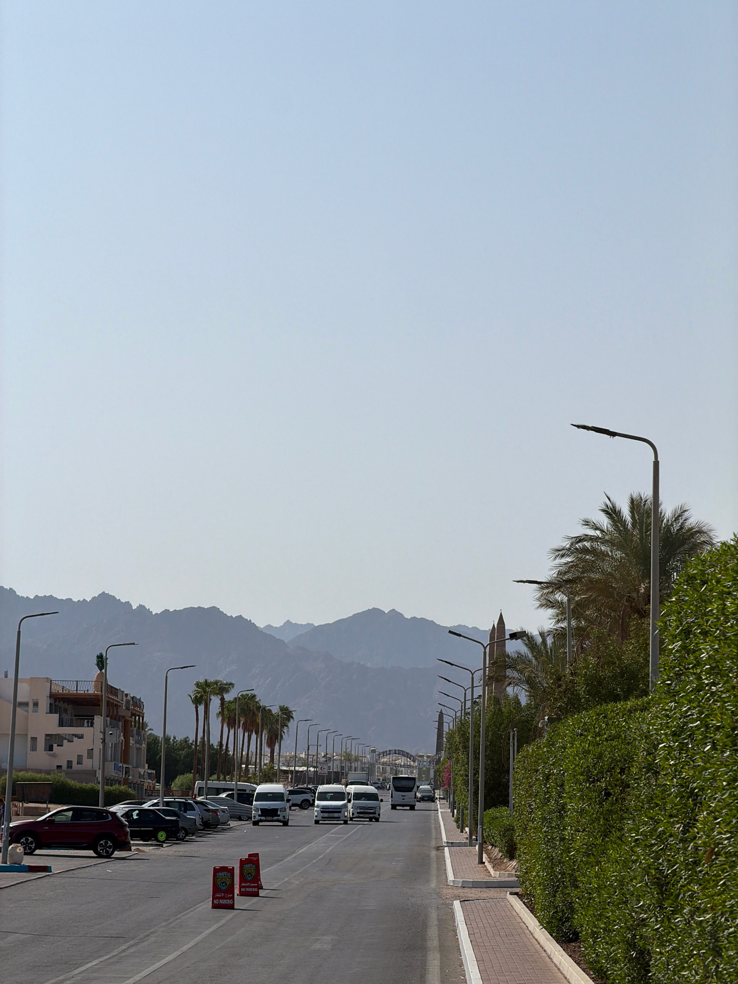 The image depicts a street scene in Sharm El Sheikh, Egypt, on a clear day with a pale blue sky. The foreground shows a paved road with several cars parked on the left side and a row of white vans and buses driving on the right side. Red and white "NO PARKING" signs are placed on the side of the road. A sidewalk with brick paving runs alongside the road, bordered by a lush green hedge. Palm trees and other vegetation line the street. In the background, a range of hazy, rocky mountains is visible. Buildings with balconies and a tower-like structure can be seen on the left side of the street. The overall impression is one of a moderately busy street in a sunny, arid environment.