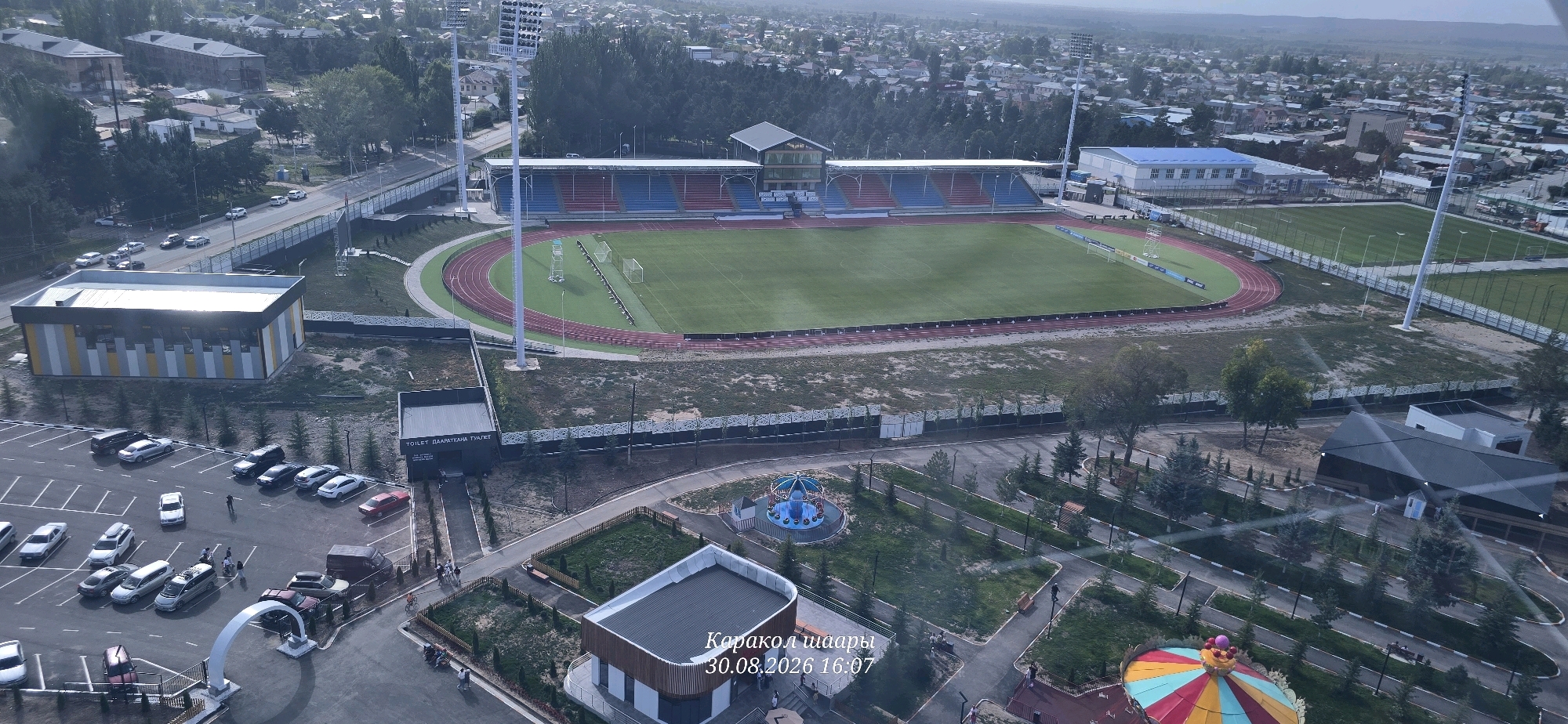 The image is an aerial view of a sports stadium and surrounding facilities in Karakol City, Kyrgyzstan. The stadium features a green football field and a red running track, with bleachers colored red and blue. Adjacent to the stadium are training fields, a large parking lot with numerous cars, and several buildings. One building is a modern, multi-colored structure. Another building is marked as a toilet facility. In the foreground, there is a park area with walking paths, benches, and a carousel. The city of Karakol is visible in the background, with residential buildings and trees. A watermark in the lower right corner reads "Каракол шаары 30.08.2026 16:07", indicating the location and date the photo was taken. There are several people visible, some walking, some in the parking lot, and some in the park area. The overall scene depicts a well-maintained sports complex within an urban environment.