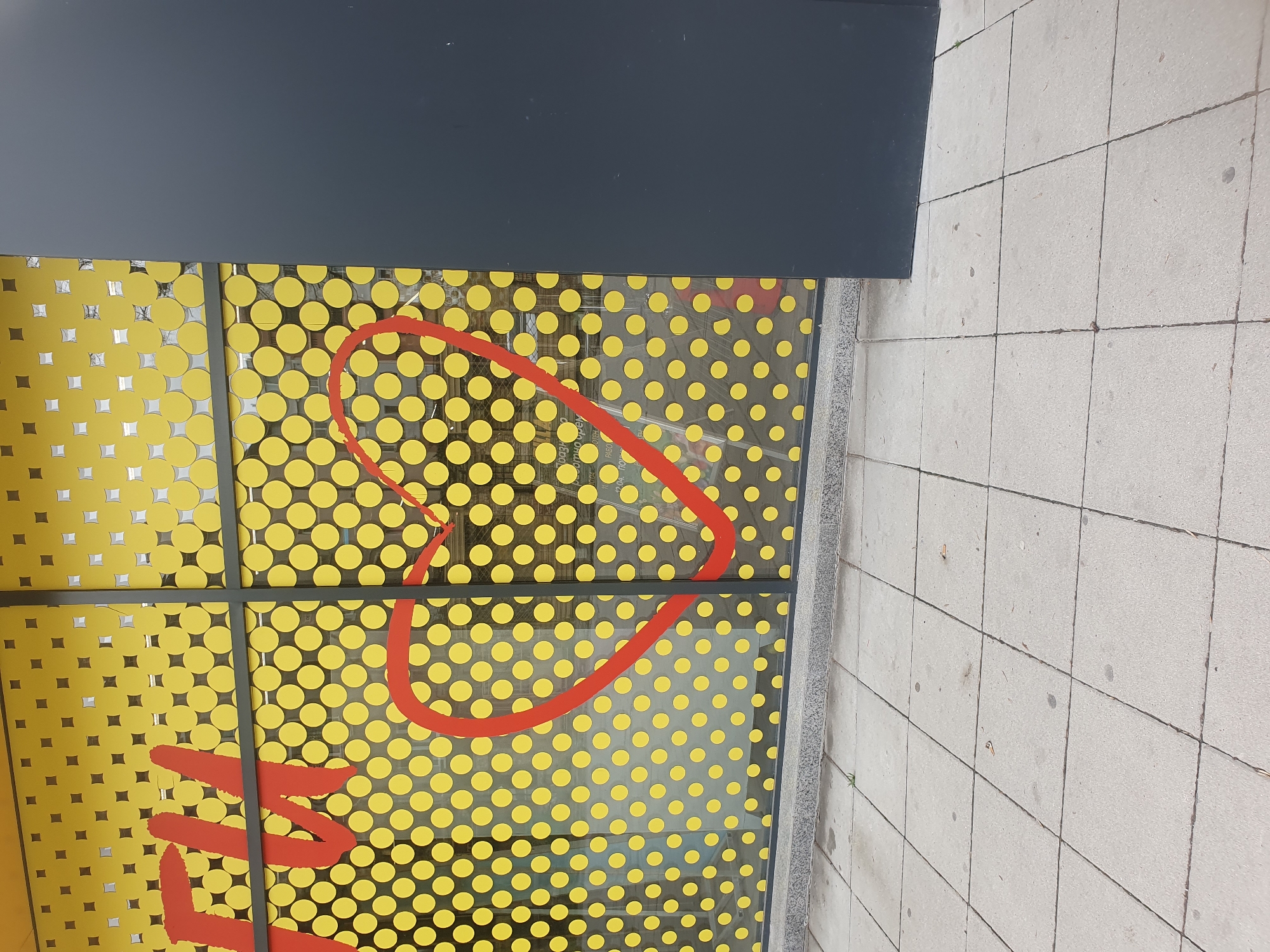 The image shows a section of a building facade in Plovdiv, Bulgaria. The facade is predominantly yellow with a pattern of evenly spaced, round yellow dots. A large red graphic, resembling an elongated heart shape, is overlaid on the yellow background. The graphic extends across multiple window panes. The windows are framed by a dark gray grid. Above the windows is a solid dark gray overhang. To the right of the facade is a section of a paved pedestrian walkway, composed of light gray square tiles. The image appears to be taken at street level.