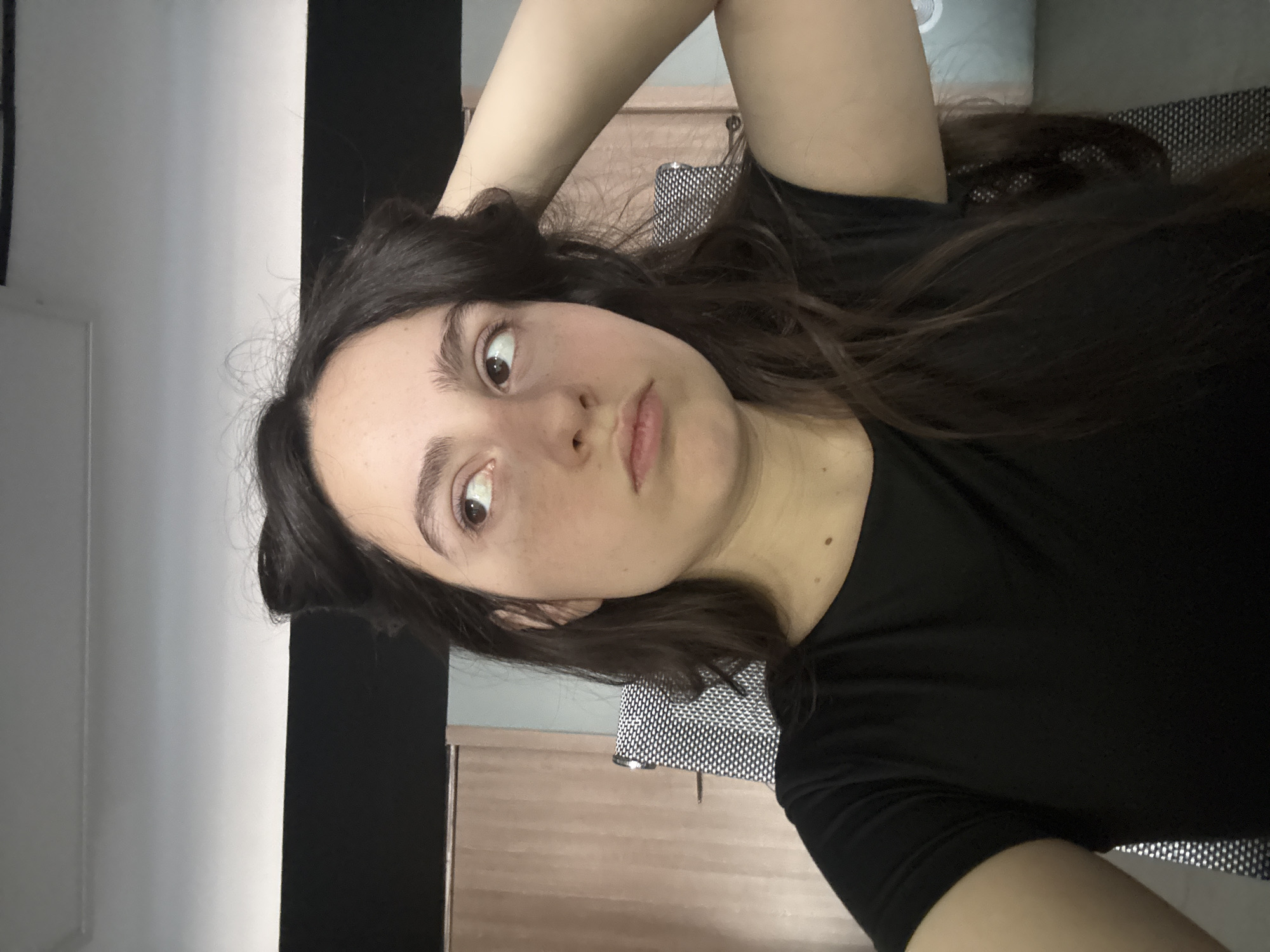 A young woman with long, dark brown hair is shown in a selfie-style photograph. She is wearing a black t-shirt. She is indoors, with a light-colored wall and dark object in the background. There is a patterned object behind her, possibly a chair. The location is indeterminate, and there is no evidence to suggest it is in Yerevan, Armenia.